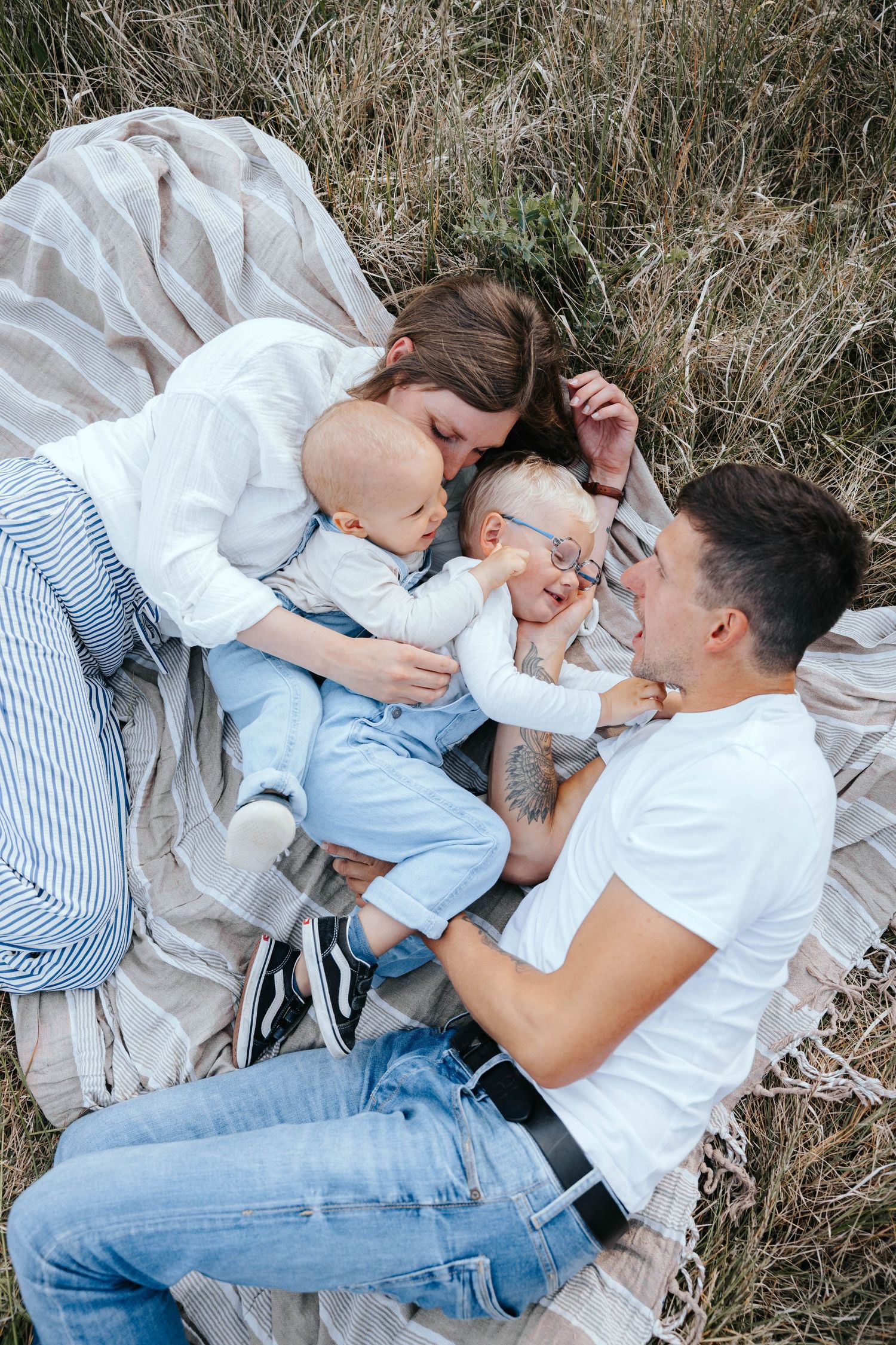 Young family with two babies lying on blanket in grass field, sharing tender moments outdoors.
