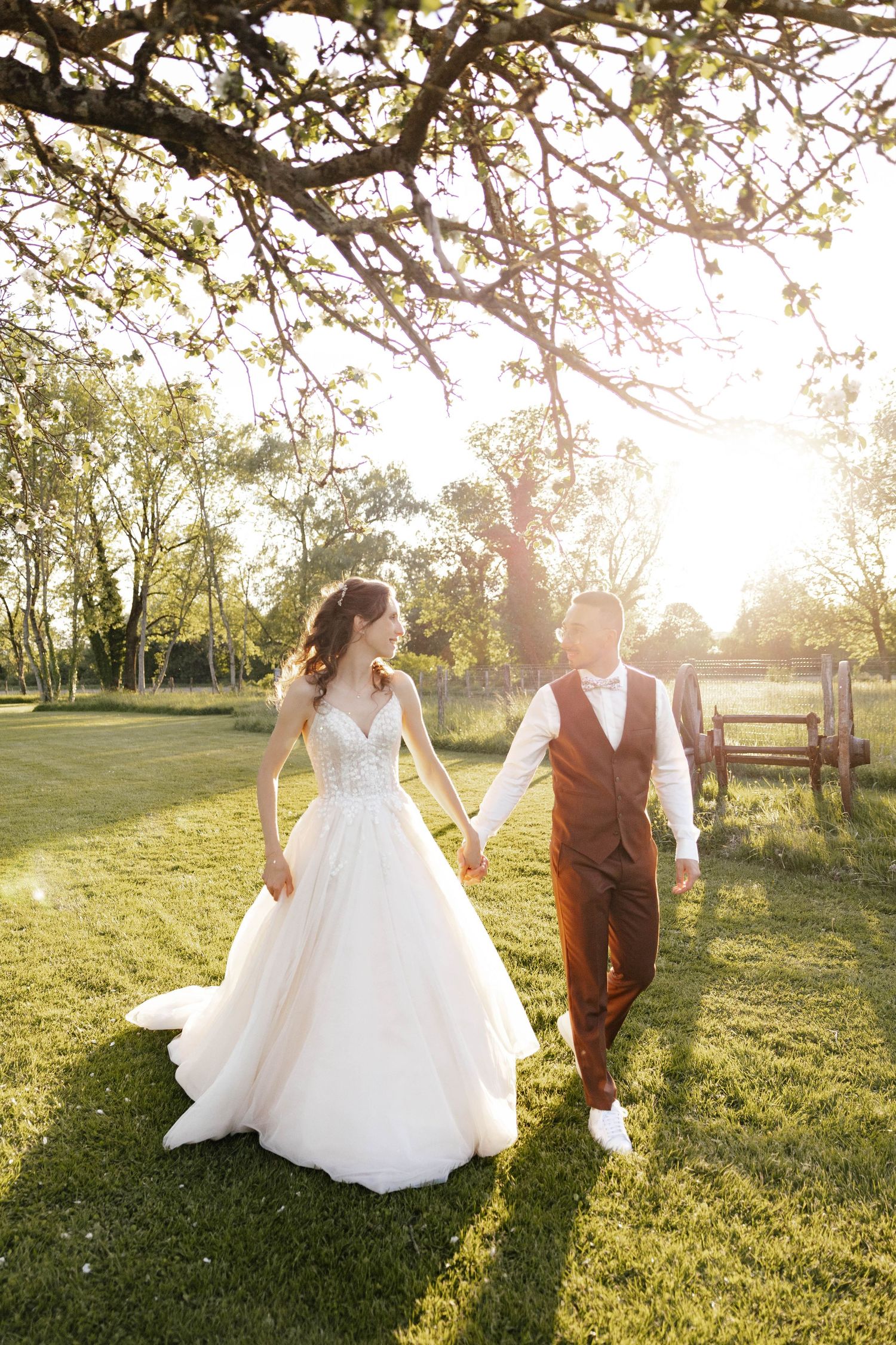 Bride and groom walking hand in hand under a blossoming tree in warm sunlight.