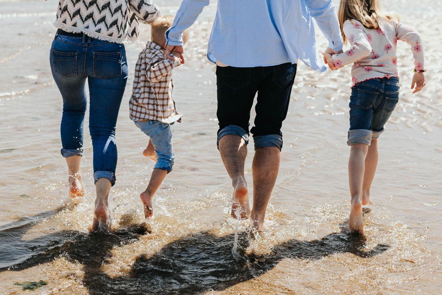 A family of four runs barefoot through shallow ocean waves, splashing water at a sunny beach.