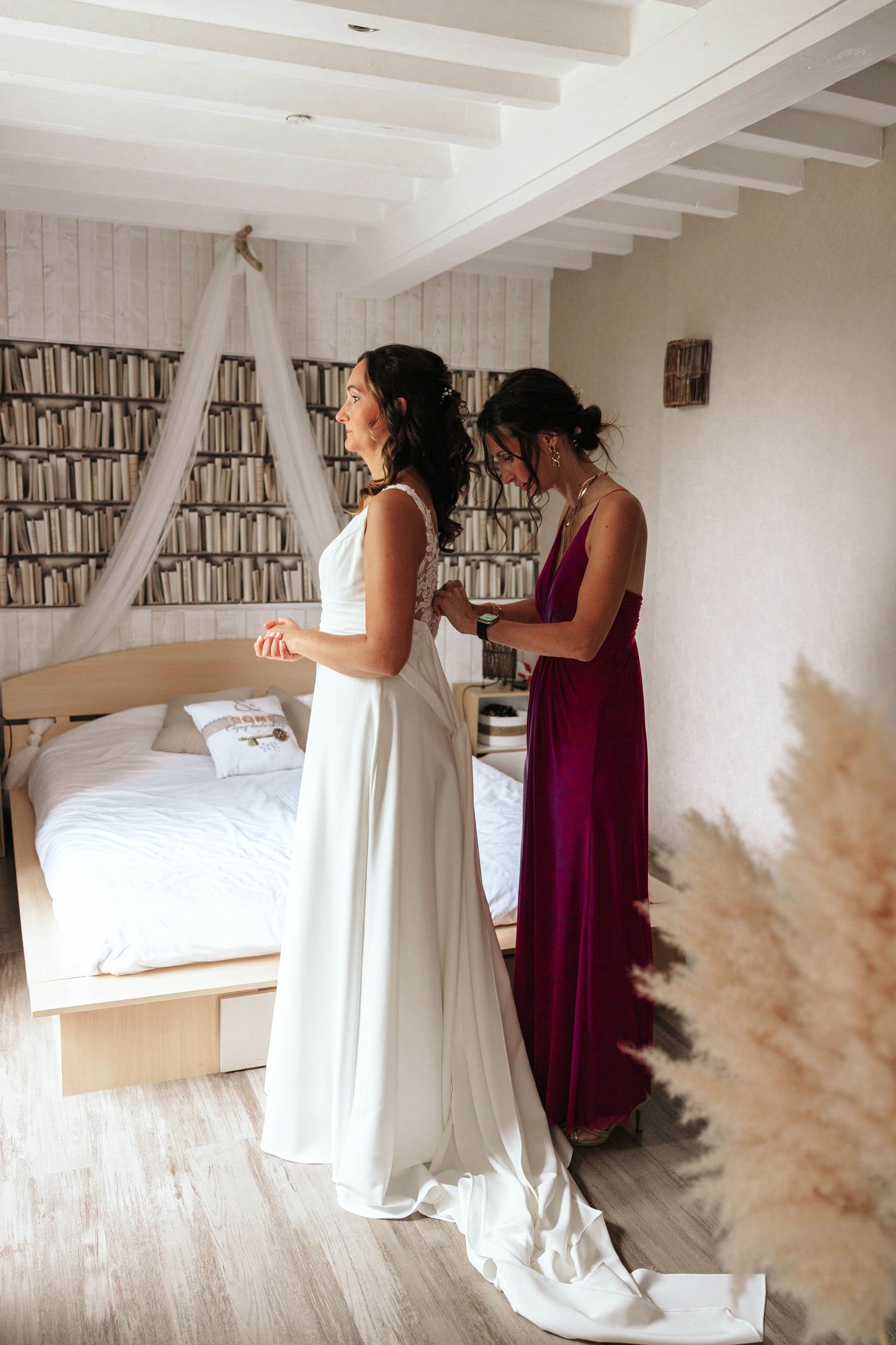 Bridesmaid in burgundy dress helping bride in white gown get ready in a cozy bedroom with bookshelf wallpaper.