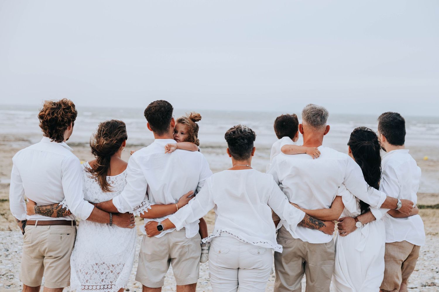 Large family in white outfits standing arm in arm with backs to camera facing the ocean.
