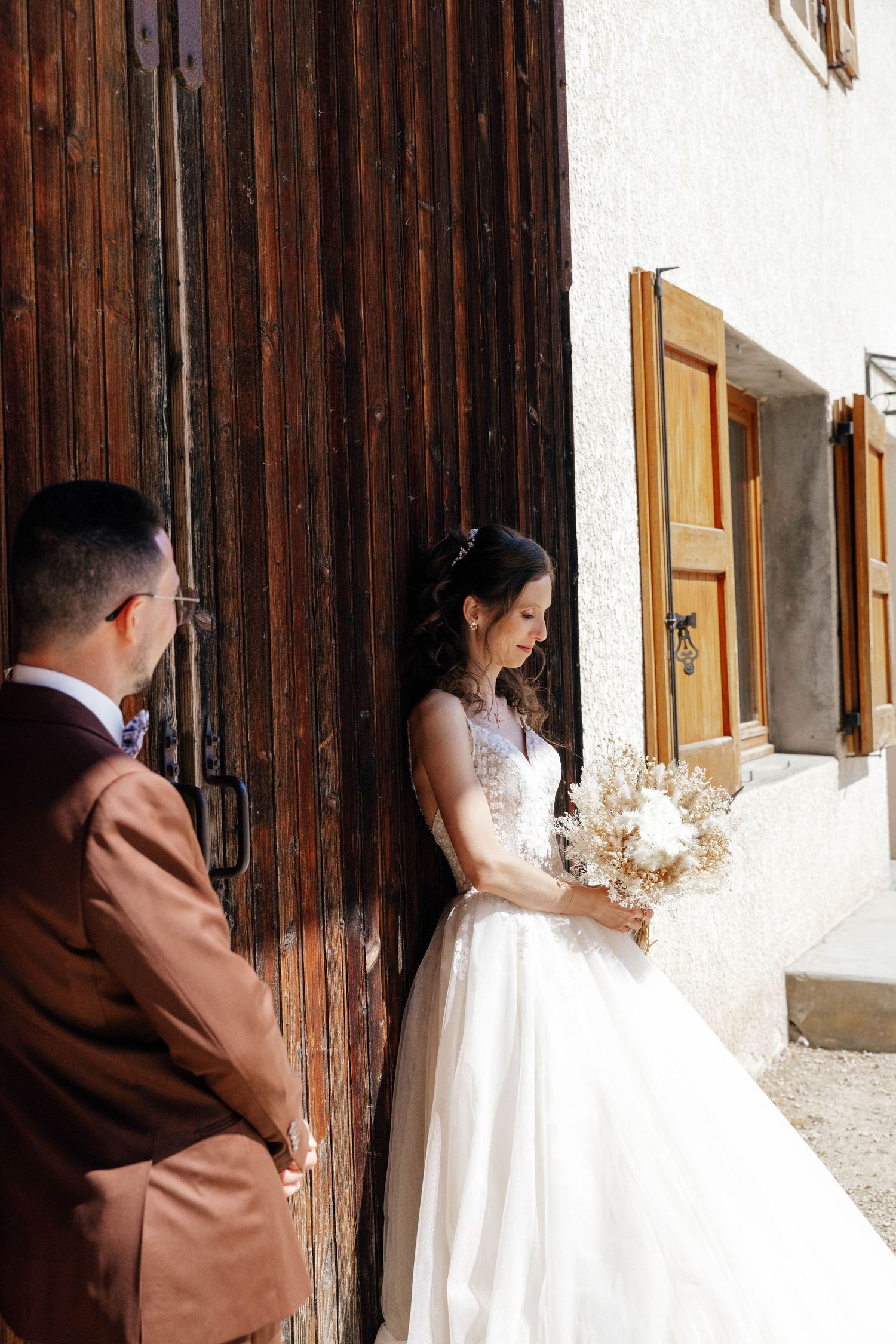Bride in white gown holding dried flower bouquet stands with groom by rustic wooden barn doors in sunlight.