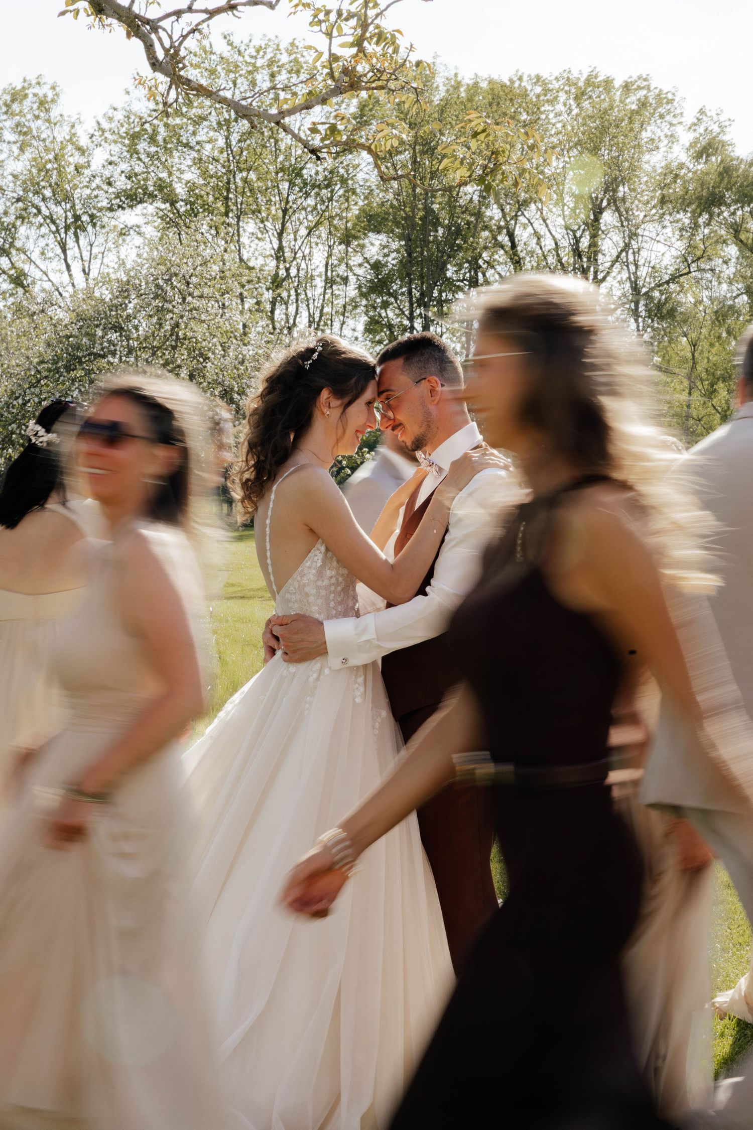 Bride and groom share a tender moment surrounded by blurred wedding guests dancing outdoors in a sunny garden setting.