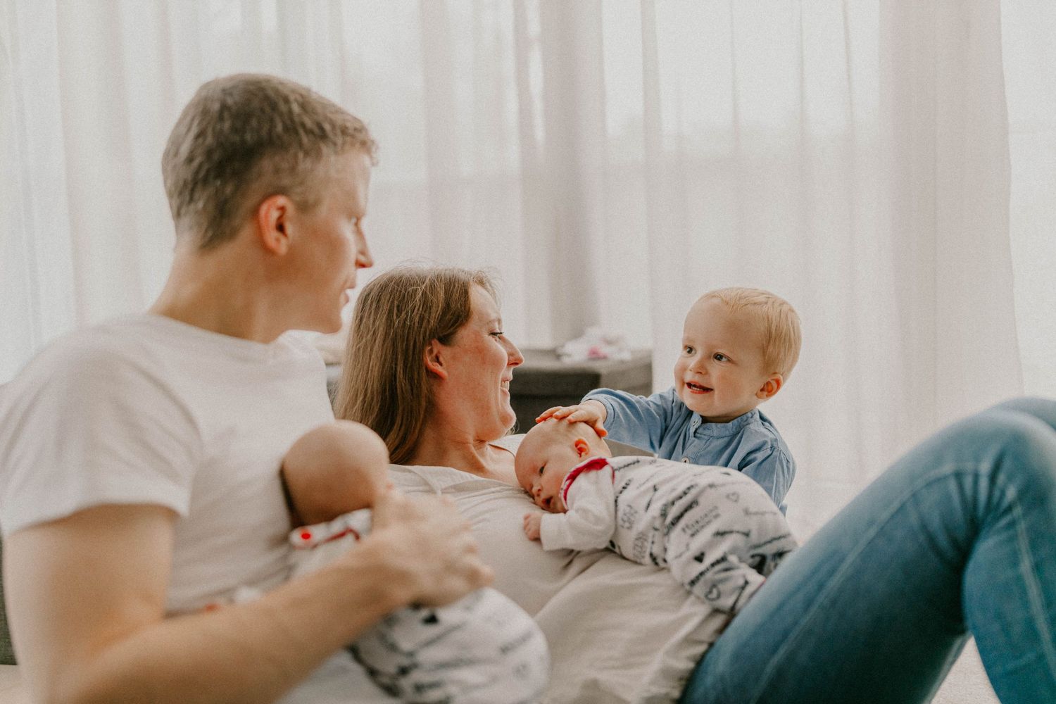 Parents relaxing on bed with newborn baby and toddler in bright white room.