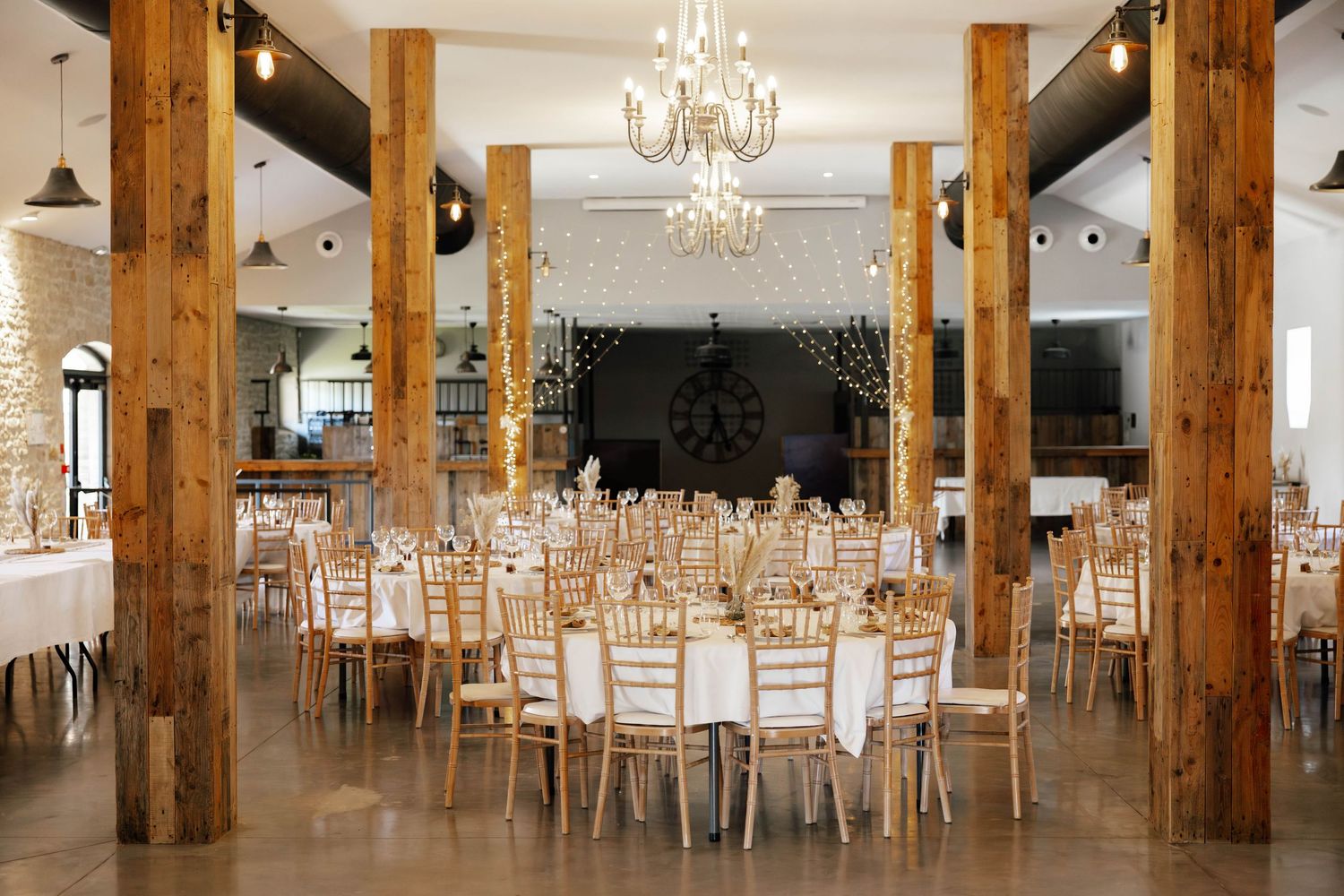Elegant wedding reception hall with rustic wood columns, gold chiavari chairs, round tables, and a chandelier.