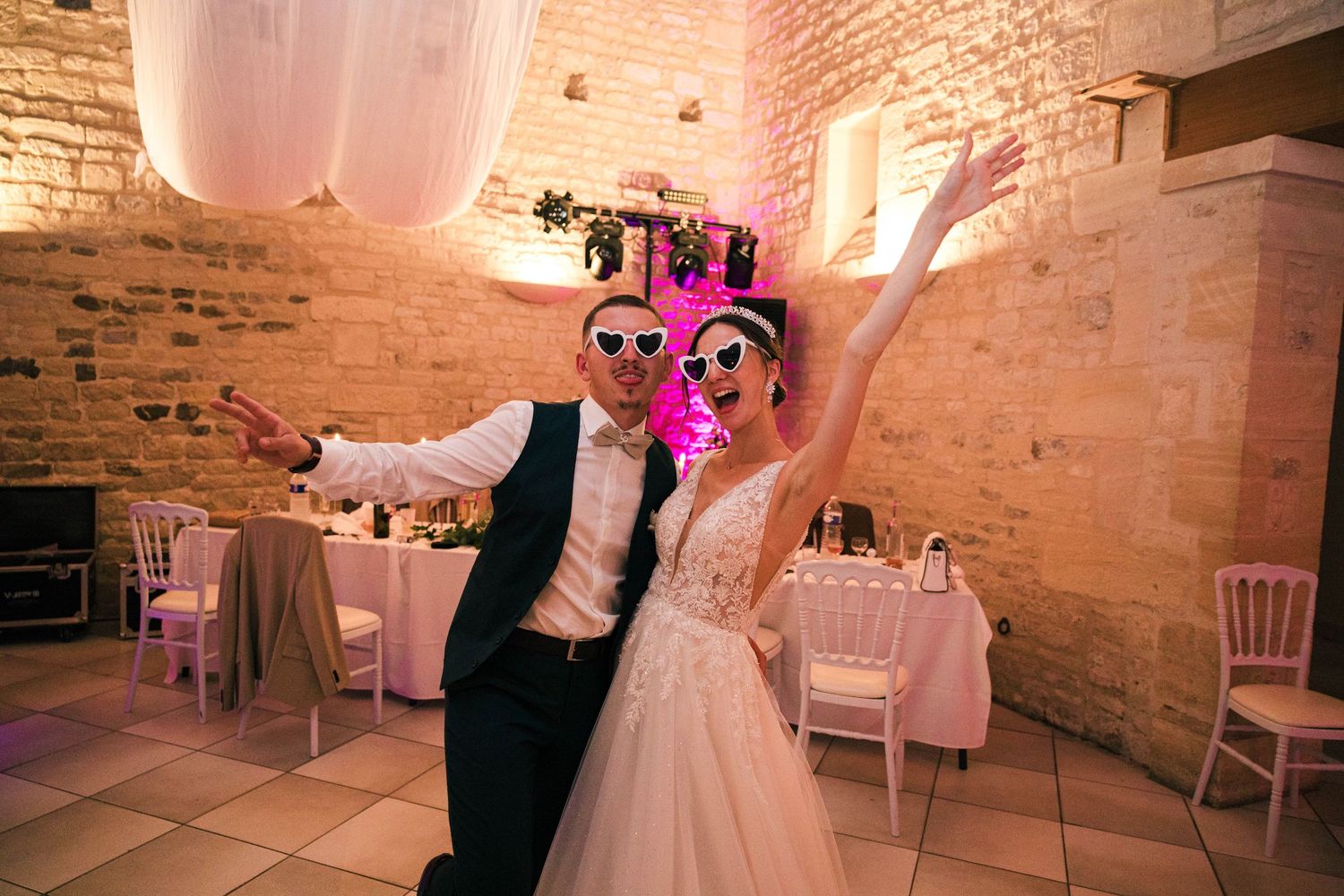 Joyful bride and groom celebrating at wedding reception wearing heart-shaped sunglasses in a rustic stone venue.