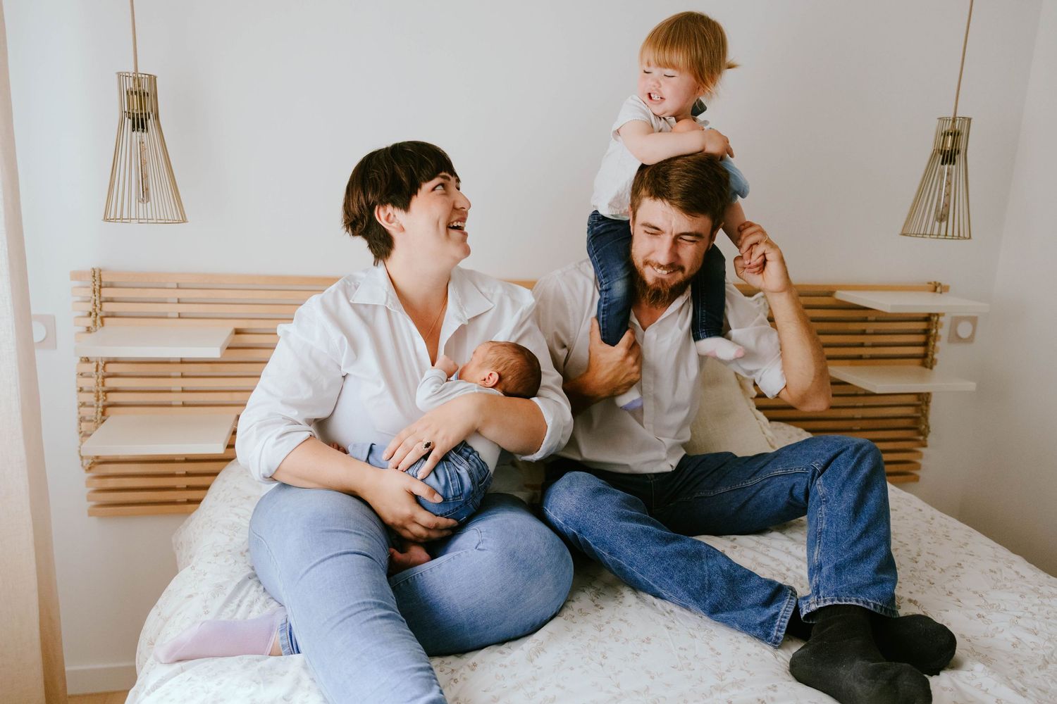 Joyful family with newborn baby and toddler on fathers shoulders laughing on white bed.