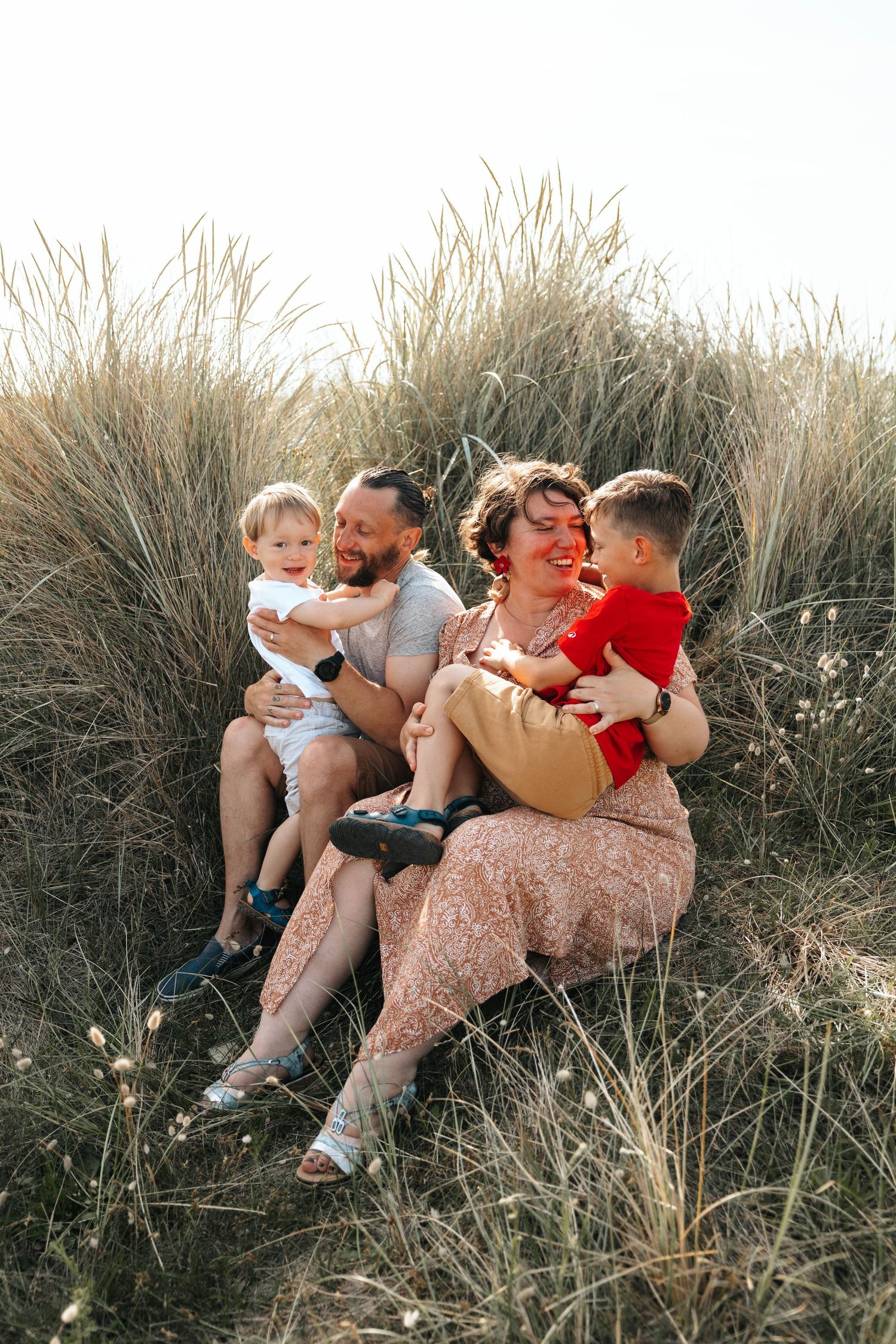 Laughing family of four sitting together in tall grass dunes, parents cuddling two young boys.