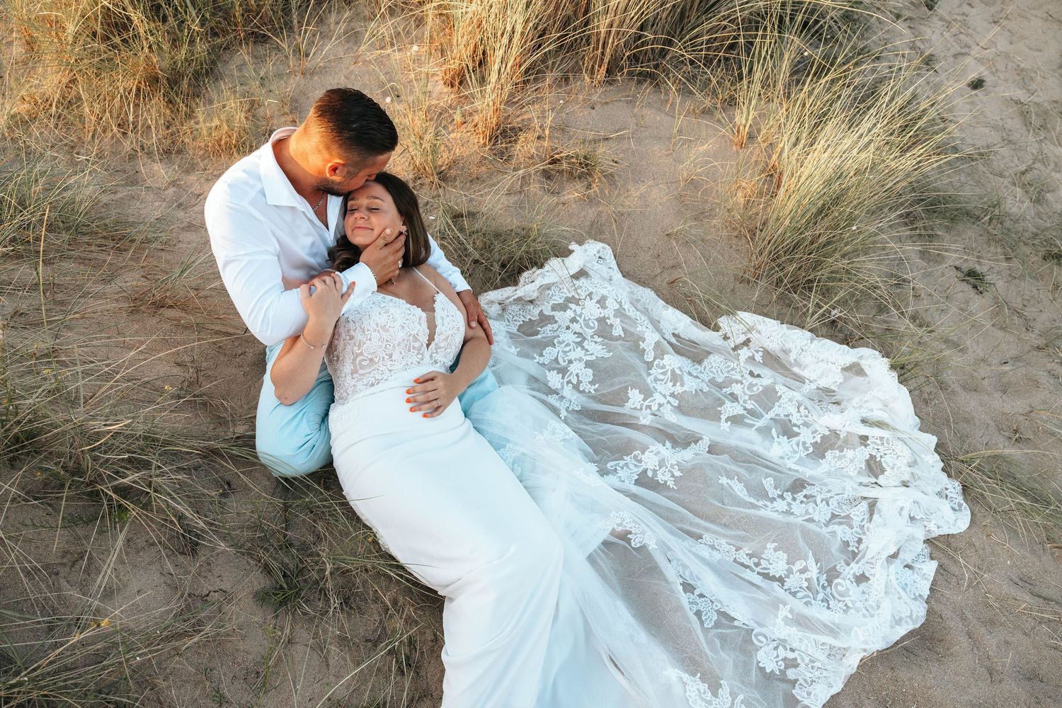 Aerial view of bride in lace gown with floral train resting against groom in sandy beach dunes at sunset.