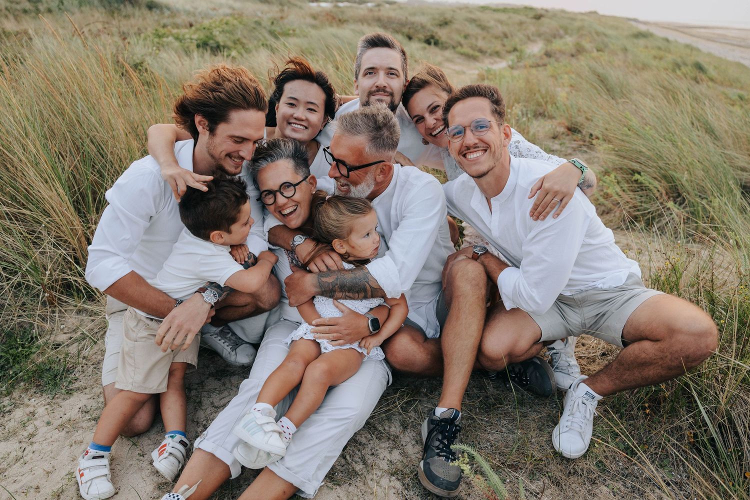 A joyful multigenerational family in white shirts cuddles together on a sandy beach dune.