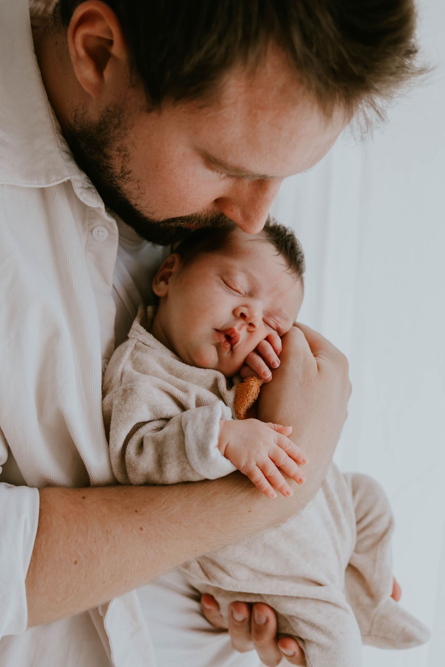 Bearded father tenderly kissing sleeping newborn baby held against his chest.