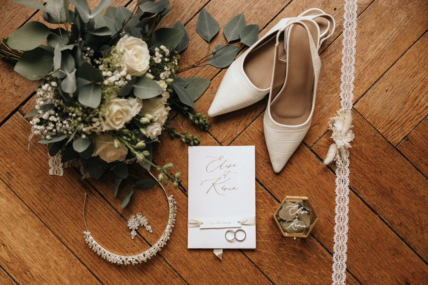 Elegant wedding flat lay with white heels, bridal bouquet, invitation, rings, and pearl jewelry on wood floor.