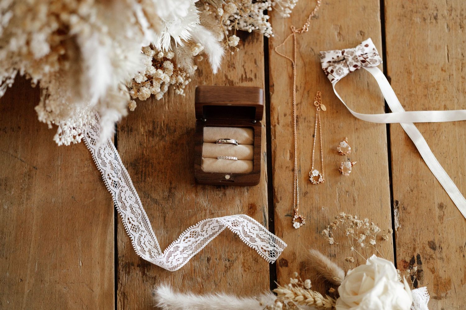 Elegant wedding flat lay with gold rings in wooden box, lace ribbon, dried flowers, and delicate jewelry on rustic wood.
