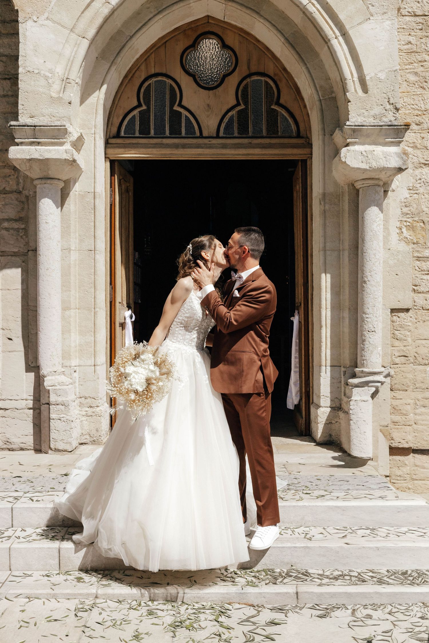Newlyweds share a kiss outside a stone church doorway, bride in white gown holding a bouquet.