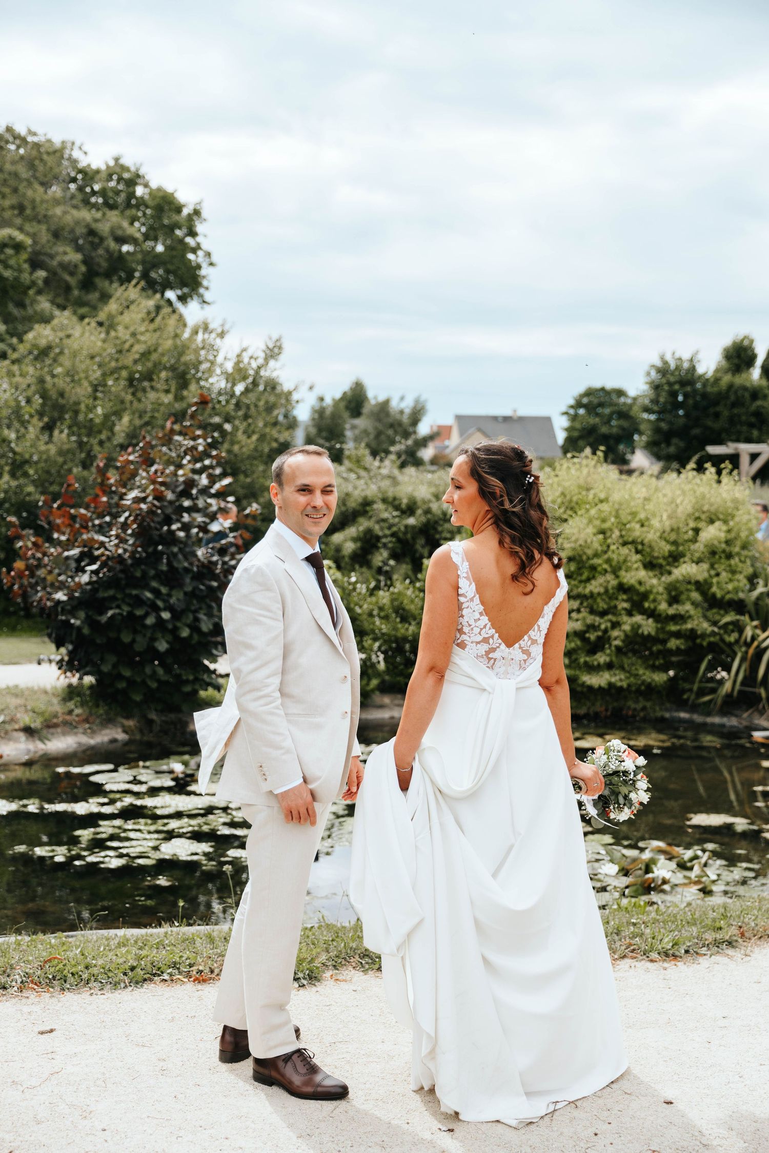 Groom smiles at camera while bride shows off her lace back wedding dress near a scenic pond.