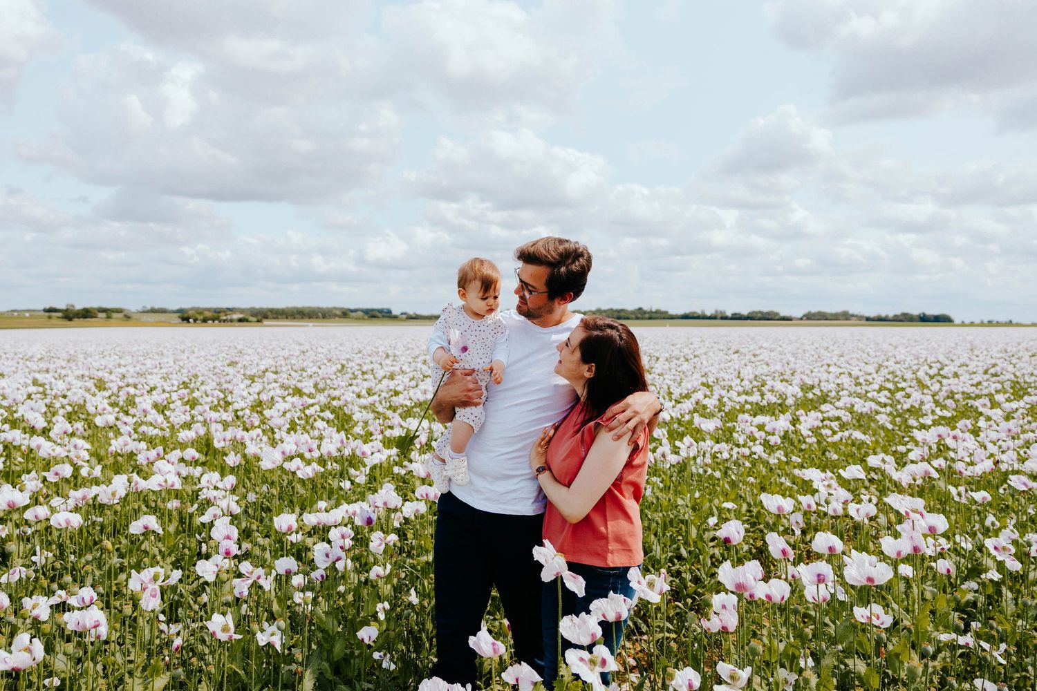 Young family with baby standing together in a vast blooming poppy flower field outdoors.