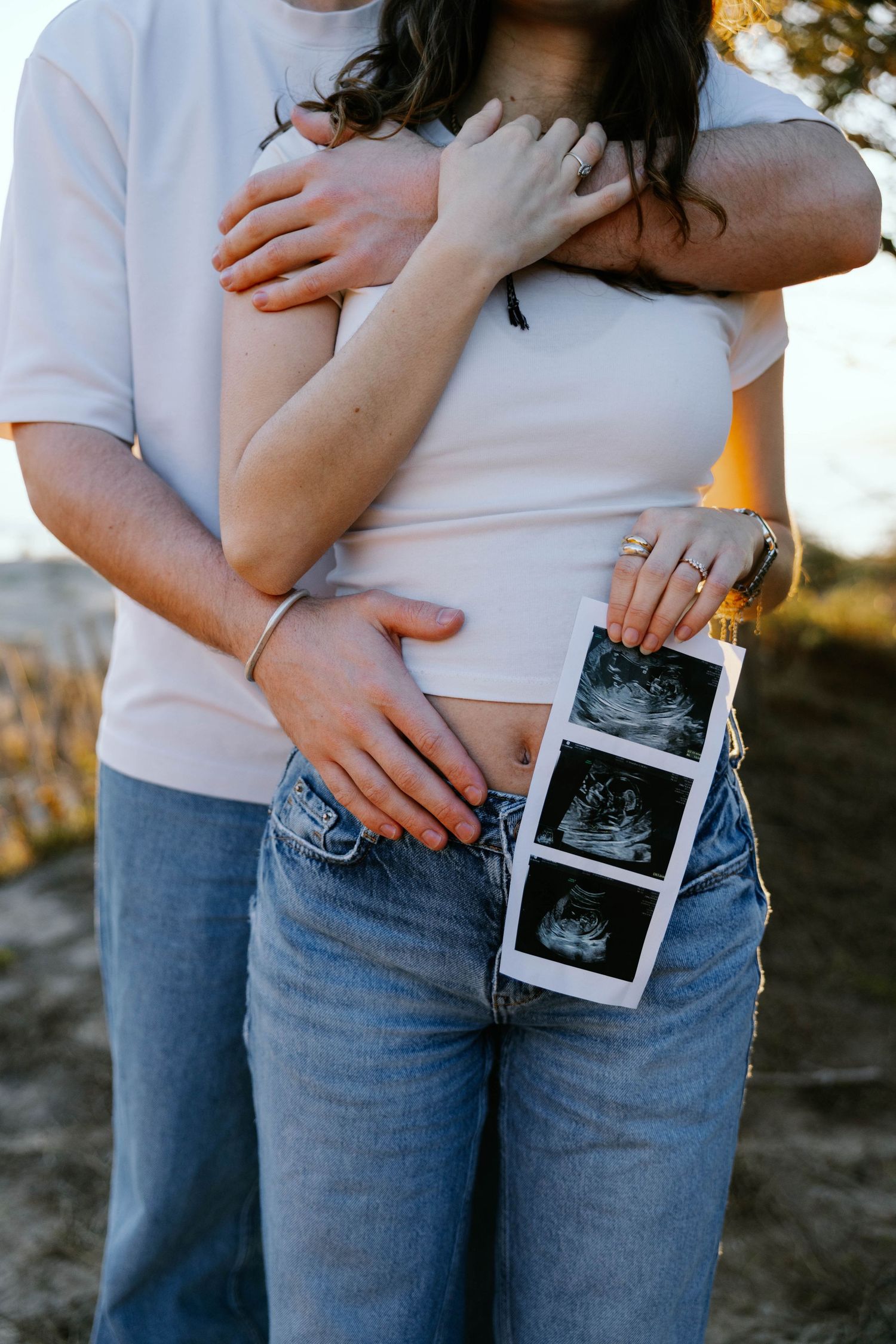 Couple embracing outdoors holding ultrasound photos, pregnancy announcement with woman in white crop top and jeans.