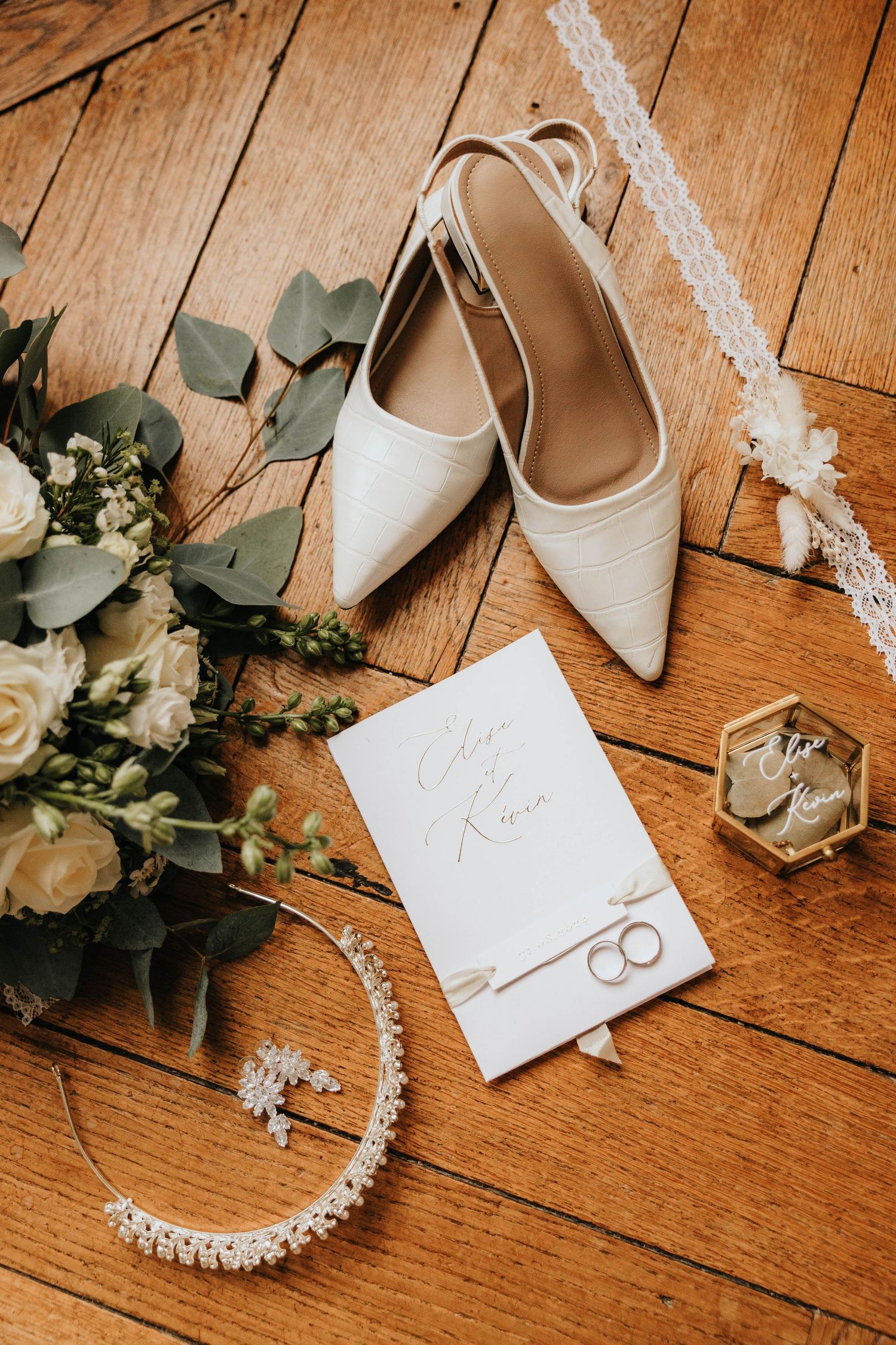Elegant bridal flat lay with white heels, floral bouquet, wedding rings, pearl necklace, and calligraphy invitation on wood floor.