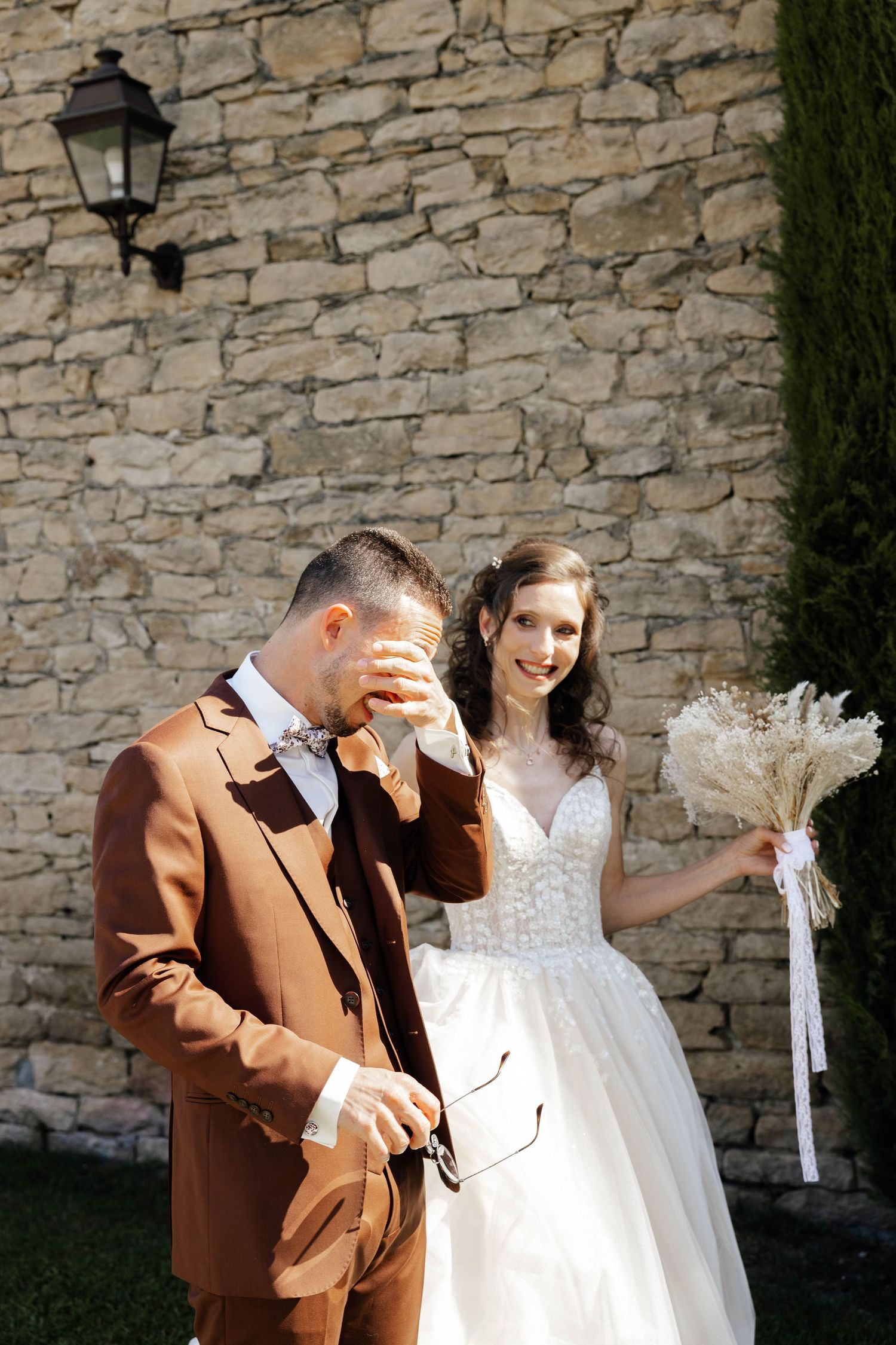 Emotional groom in brown suit wiping tears as smiling bride holds bouquet before stone wall.