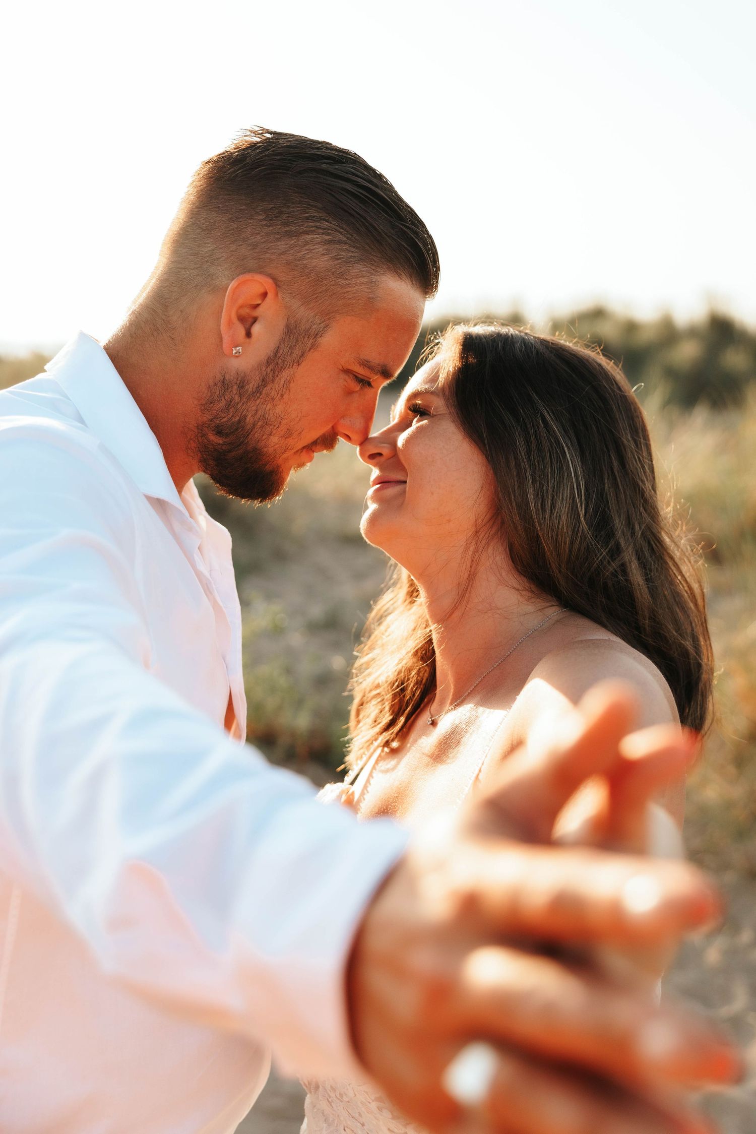 Romantic close-up of smiling bride and groom holding hands and touching foreheads in golden sunset light outdoors.