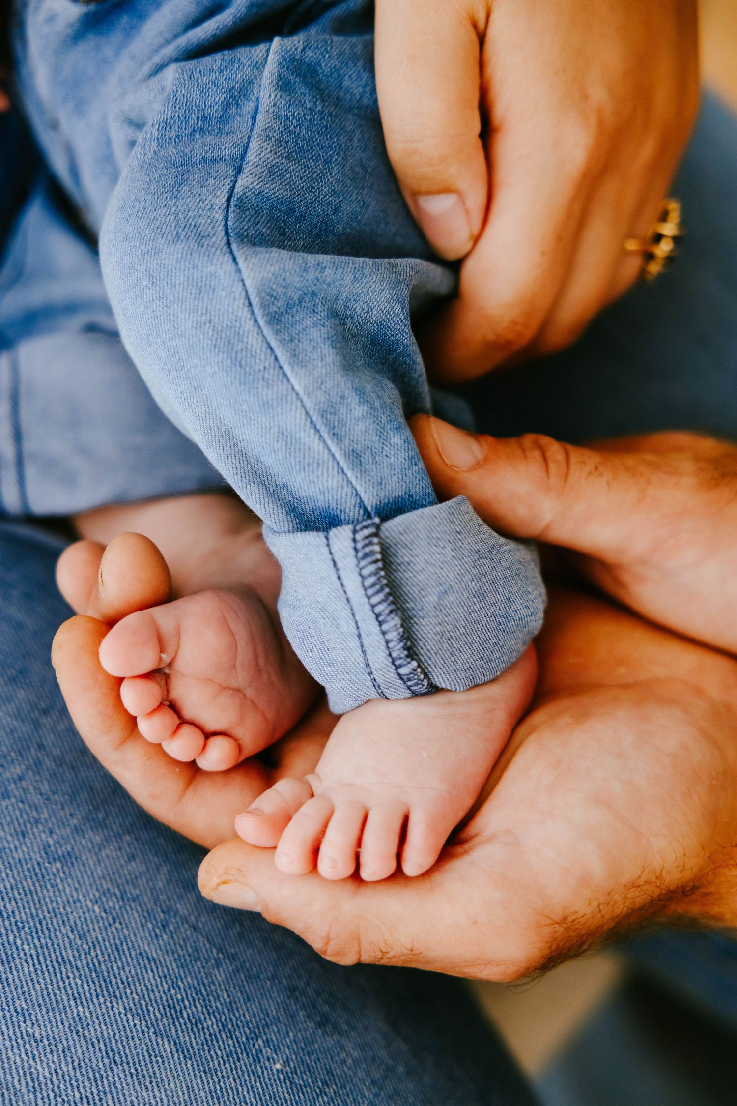 Close-up of newborn baby tiny feet cradled gently in parents hands wearing blue denim outfit.