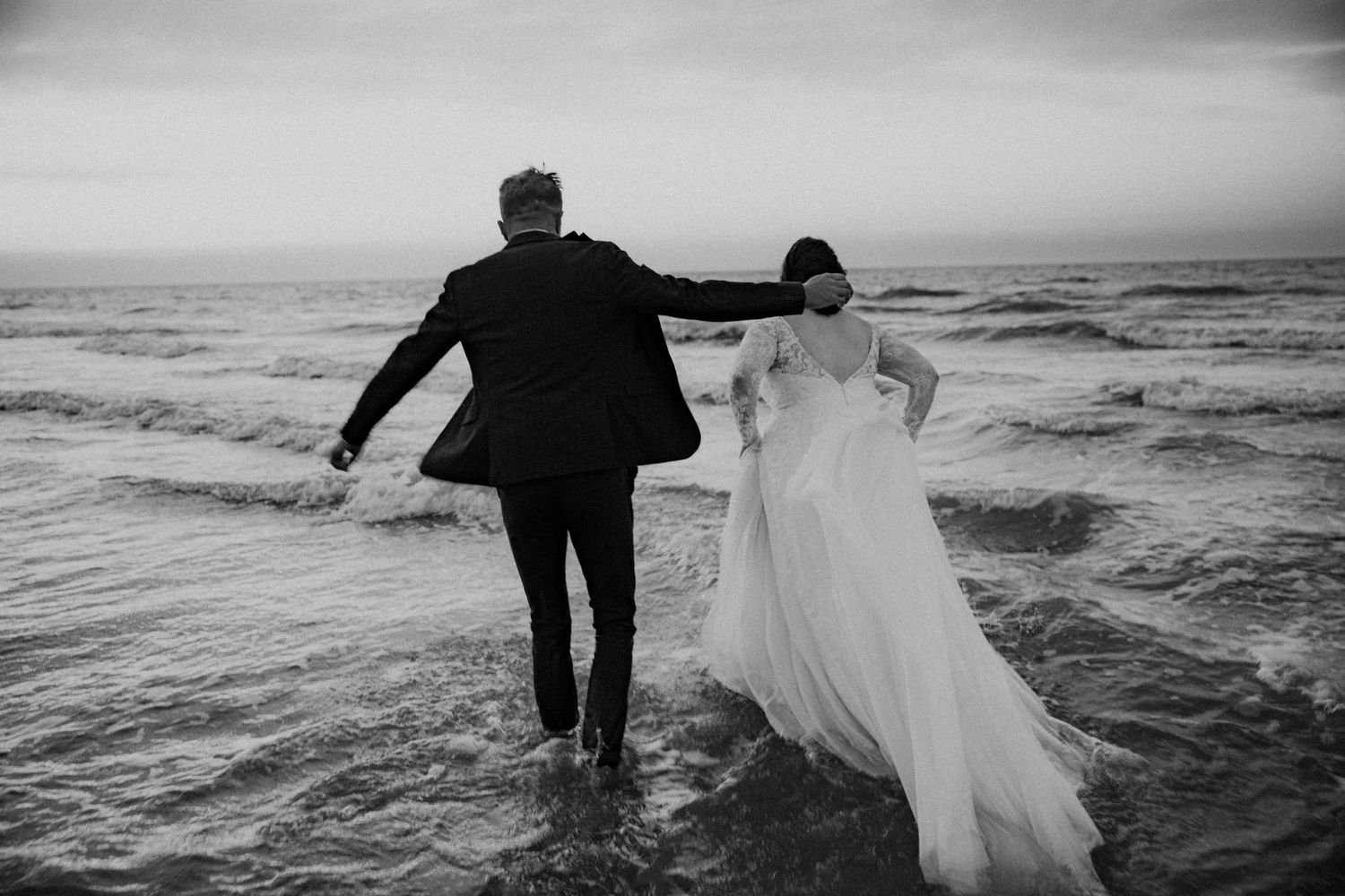 Black and white photo of bride and groom wading in ocean waves on their wedding day.