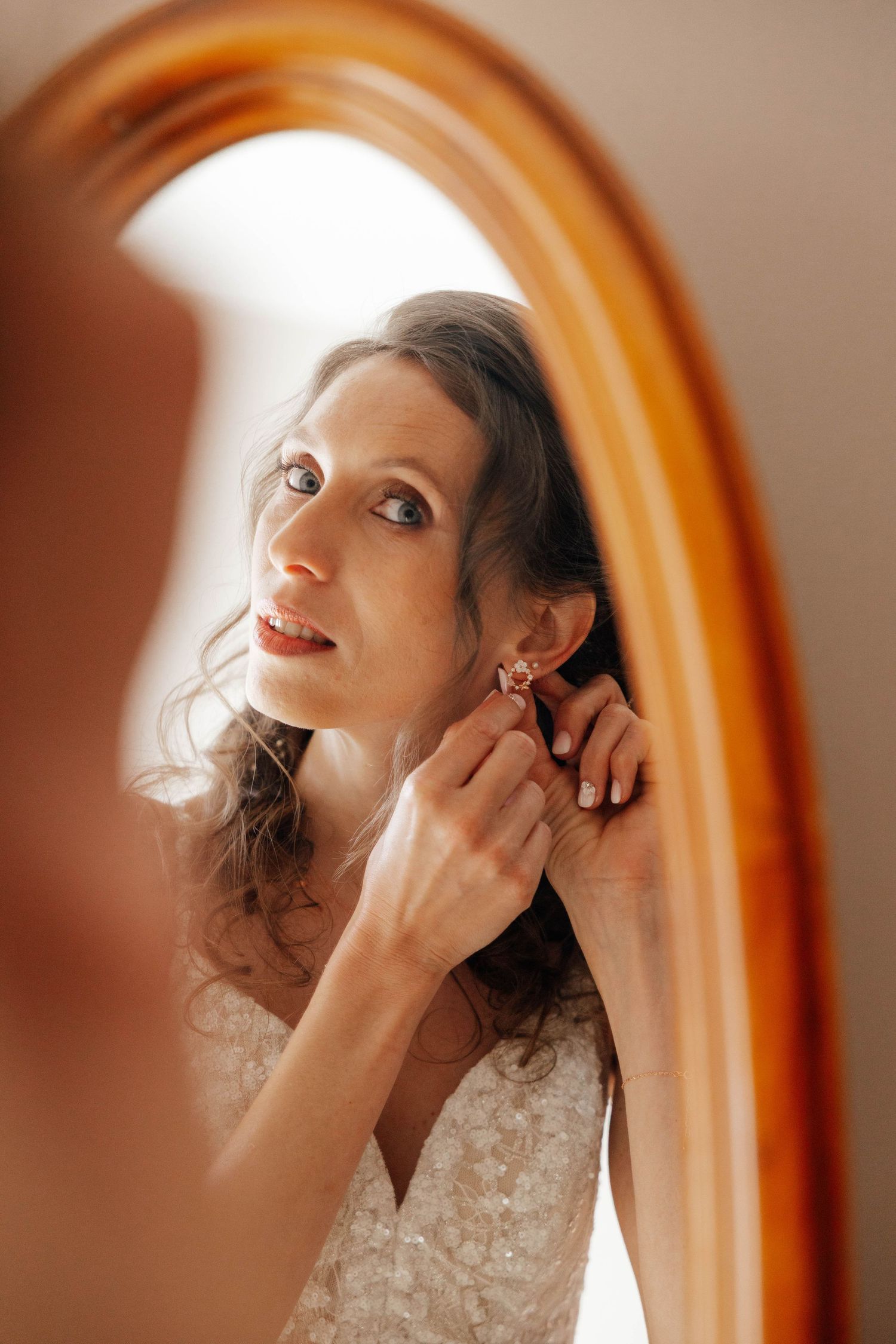 Bride putting on earrings while looking at her reflection in an oval wooden mirror.