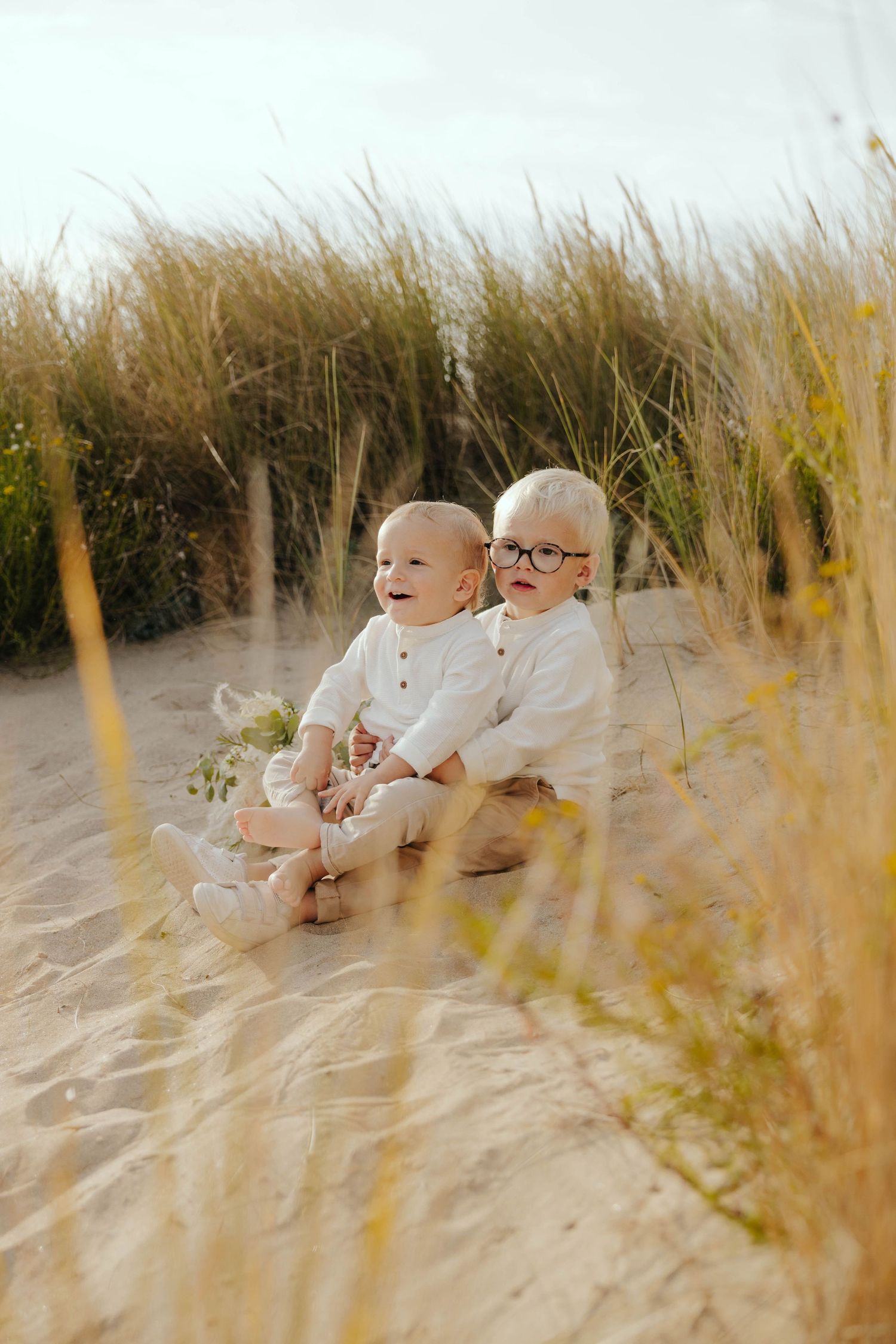 Two babies in white outfits sitting on sandy beach dunes surrounded by tall golden grass.
