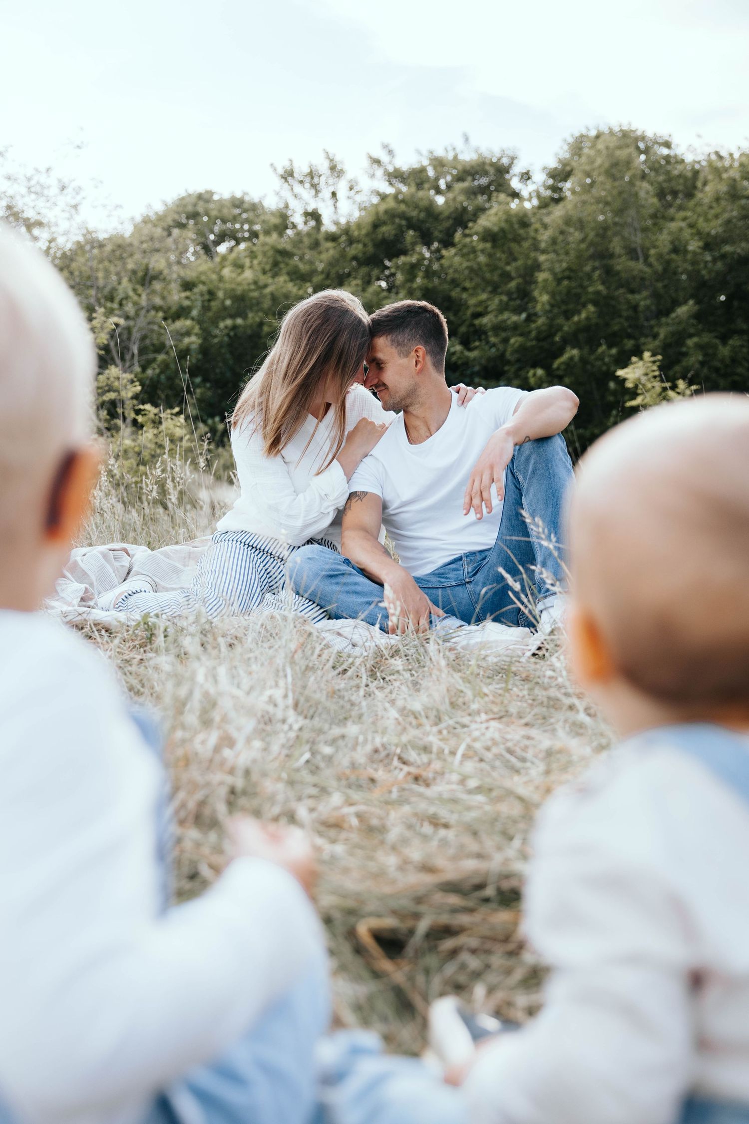 A couple sharing a romantic moment outdoors while two babies sit in the foreground on a sunny day.