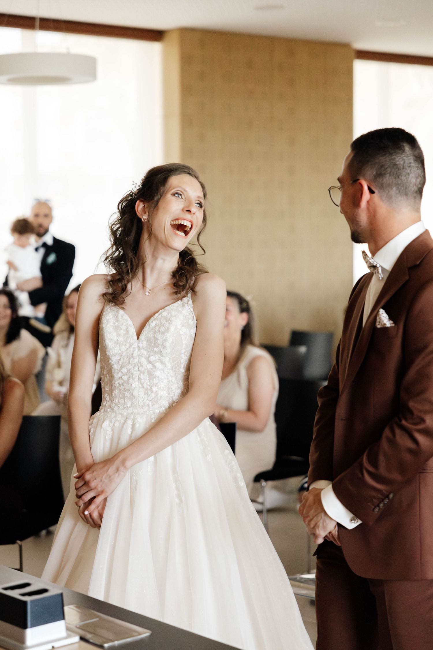 Joyful bride in lace gown laughing with groom in brown suit at wedding ceremony.