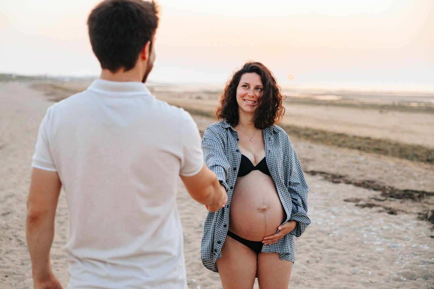 Smiling pregnant woman in denim shirt and black bikini holding her baby bump on a sandy beach at sunset.