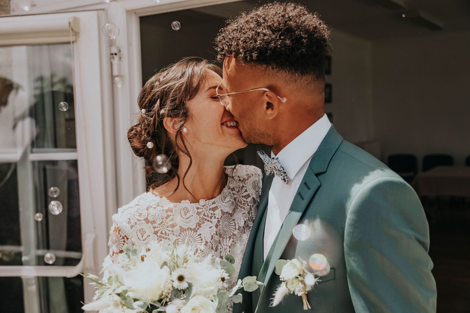 Newlywed couple sharing a romantic kiss, bride in lace gown holding white bouquet, groom in teal suit with bow tie.