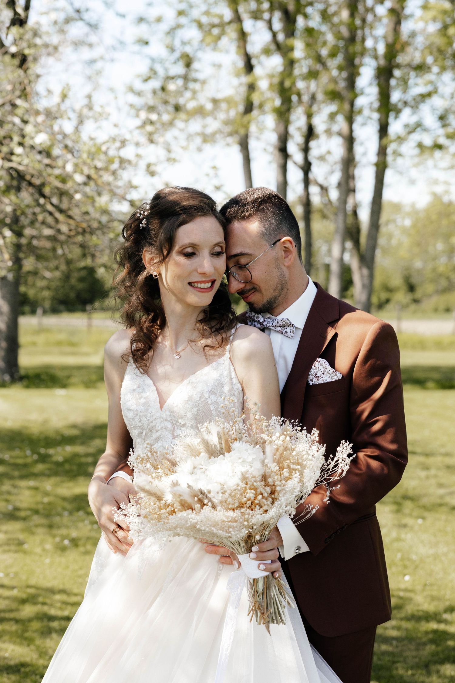 Smiling bride and groom embracing outdoors, holding elegant dried flower bouquet in sunny park.