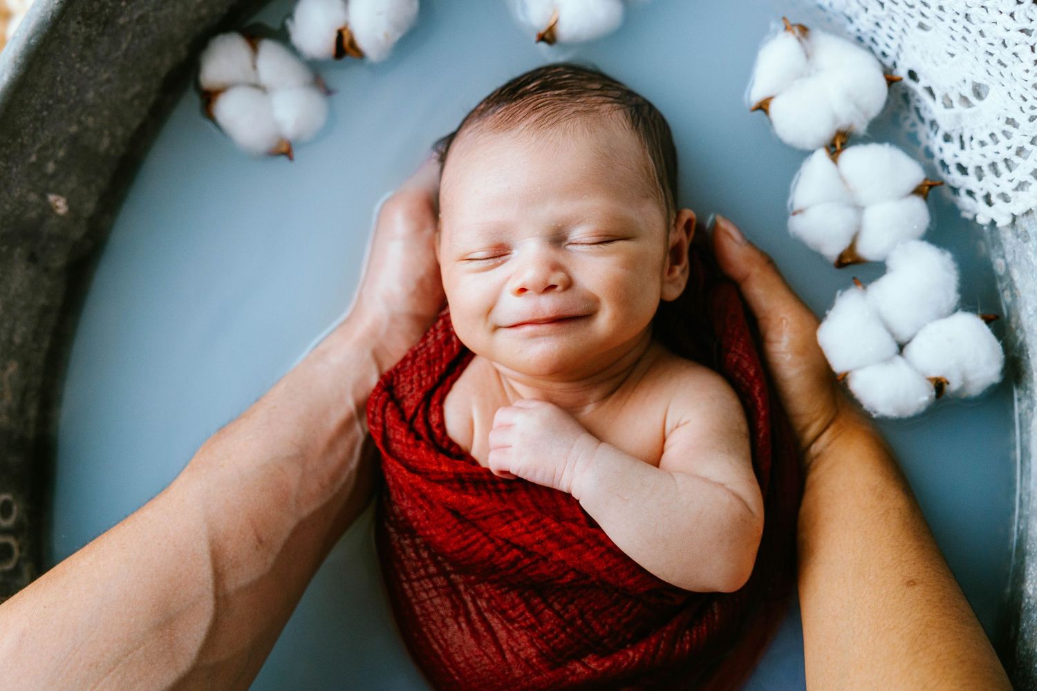 Smiling newborn baby wrapped in red knit blanket in water with cotton flowers.