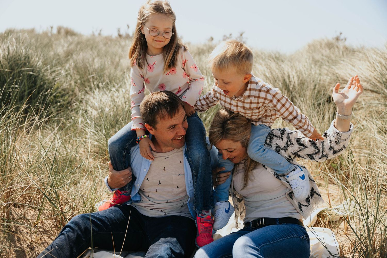 A happy family of four laughs and cuddles together among tall grass on sunny sand dunes at the beach.