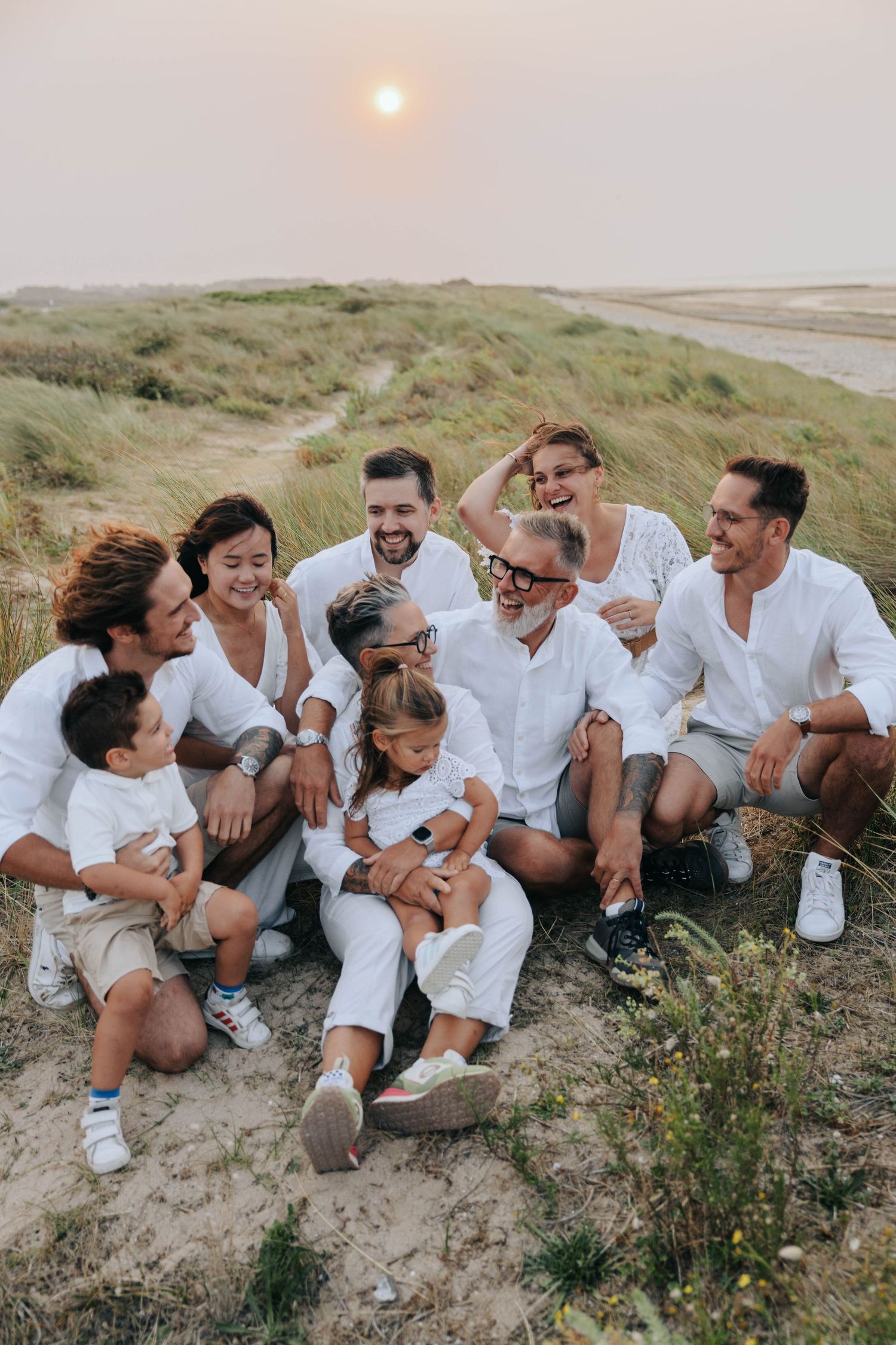 Large multigenerational family in white outfits laughing together on grassy dunes at sunset.
