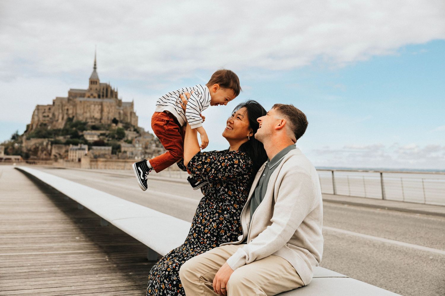 Happy family with toddler sitting on bridge with Mont Saint-Michel in background.