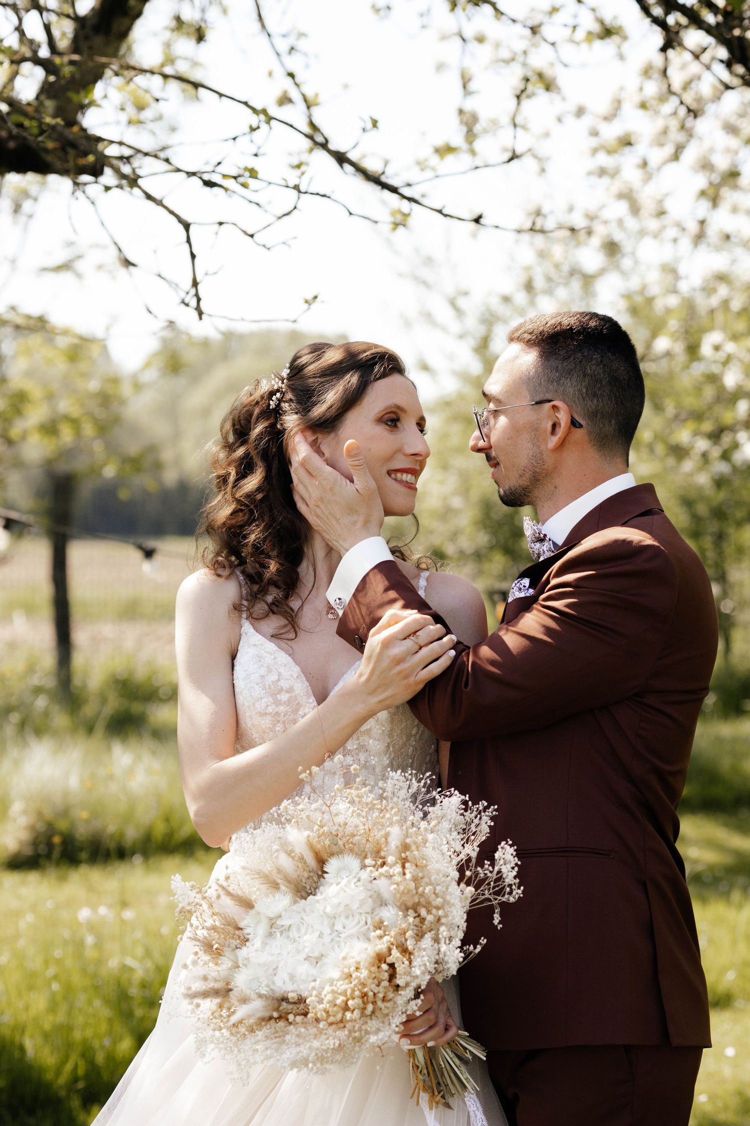 Bride and groom share tender moment outdoors, groom cupping brides face, holding dried bouquet.