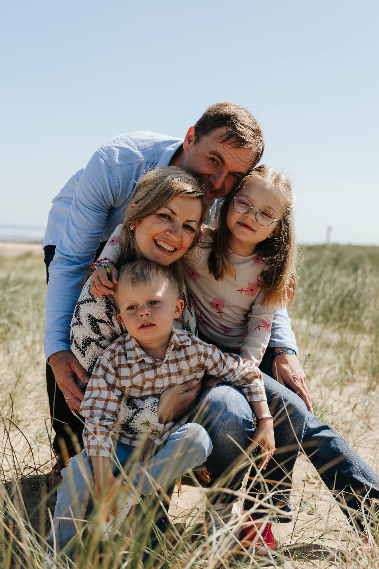 A happy family of four poses together in beach dunes, parents embracing two young children outdoors.