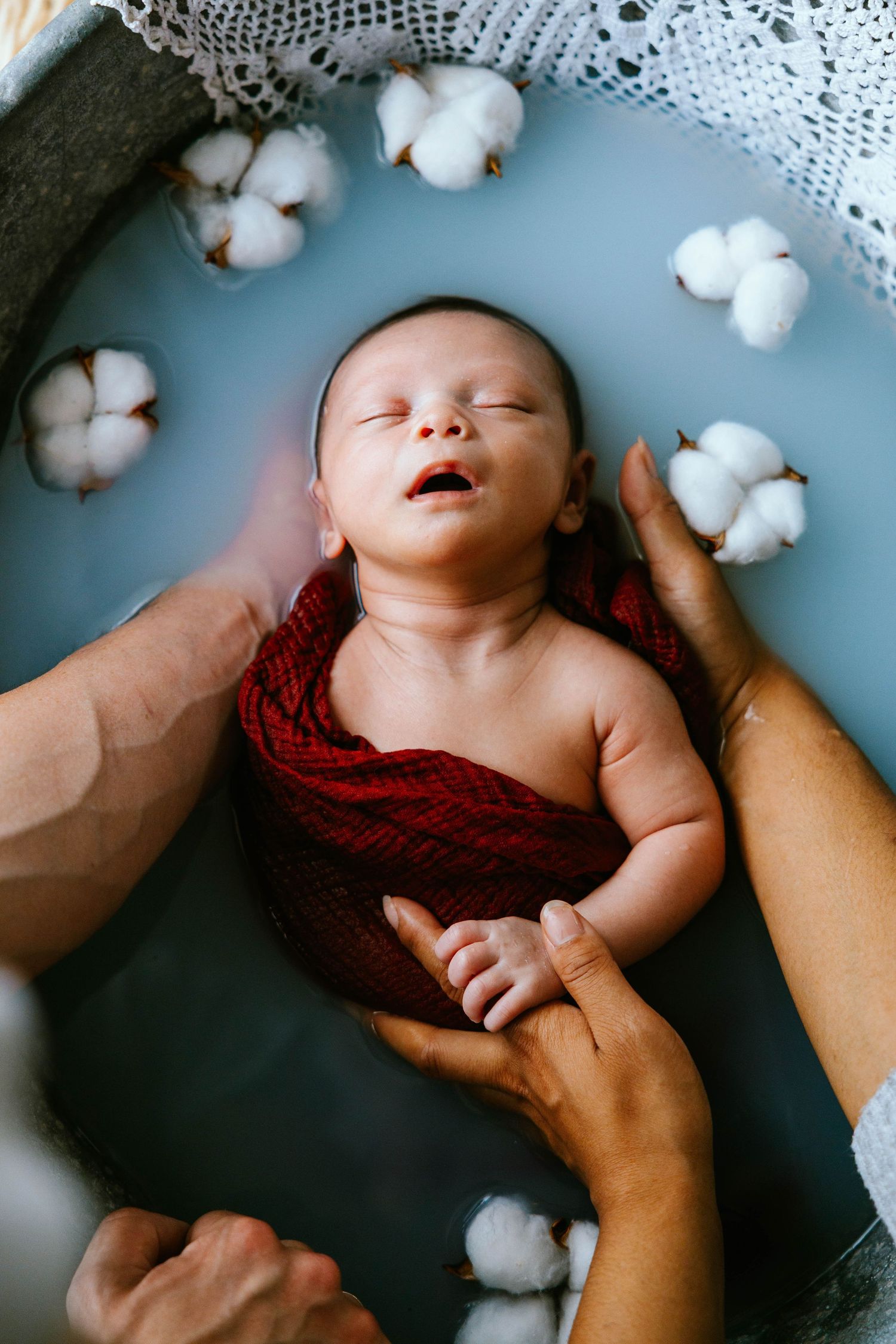 Newborn baby with open mouth wrapped in red cloth in water with cotton blooms.