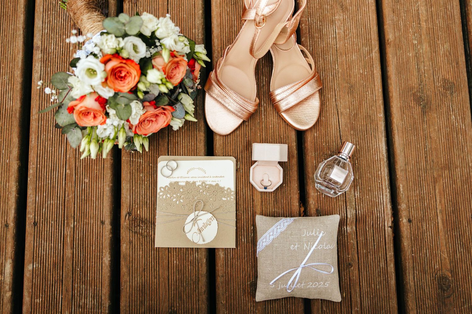 Elegant wedding flat lay with rose gold heels, bridal bouquet, invitation, ring box, and perfume on wooden deck.