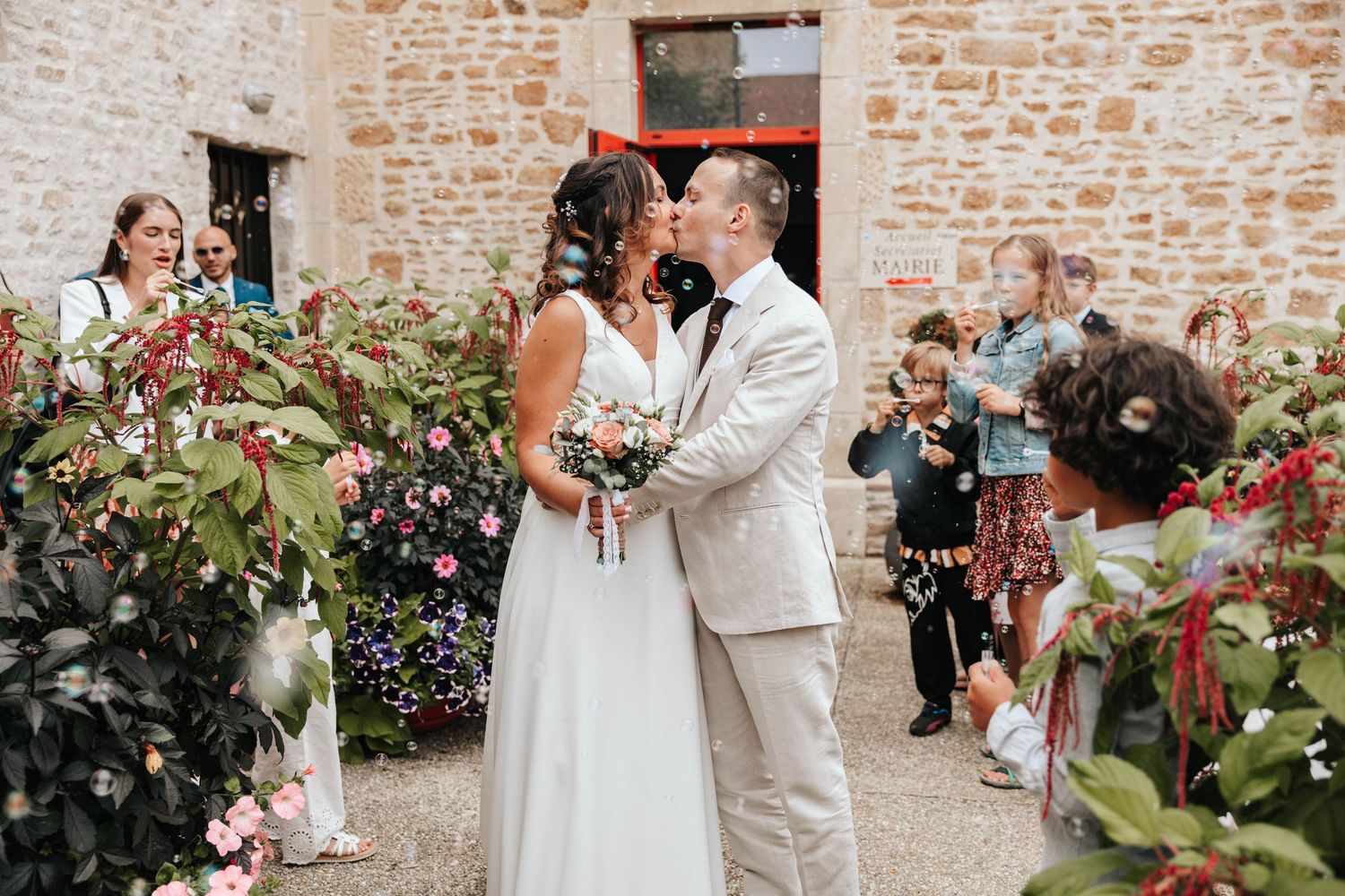 Newlyweds share a kiss outside a stone building as wedding guests celebrate around them with flowers.