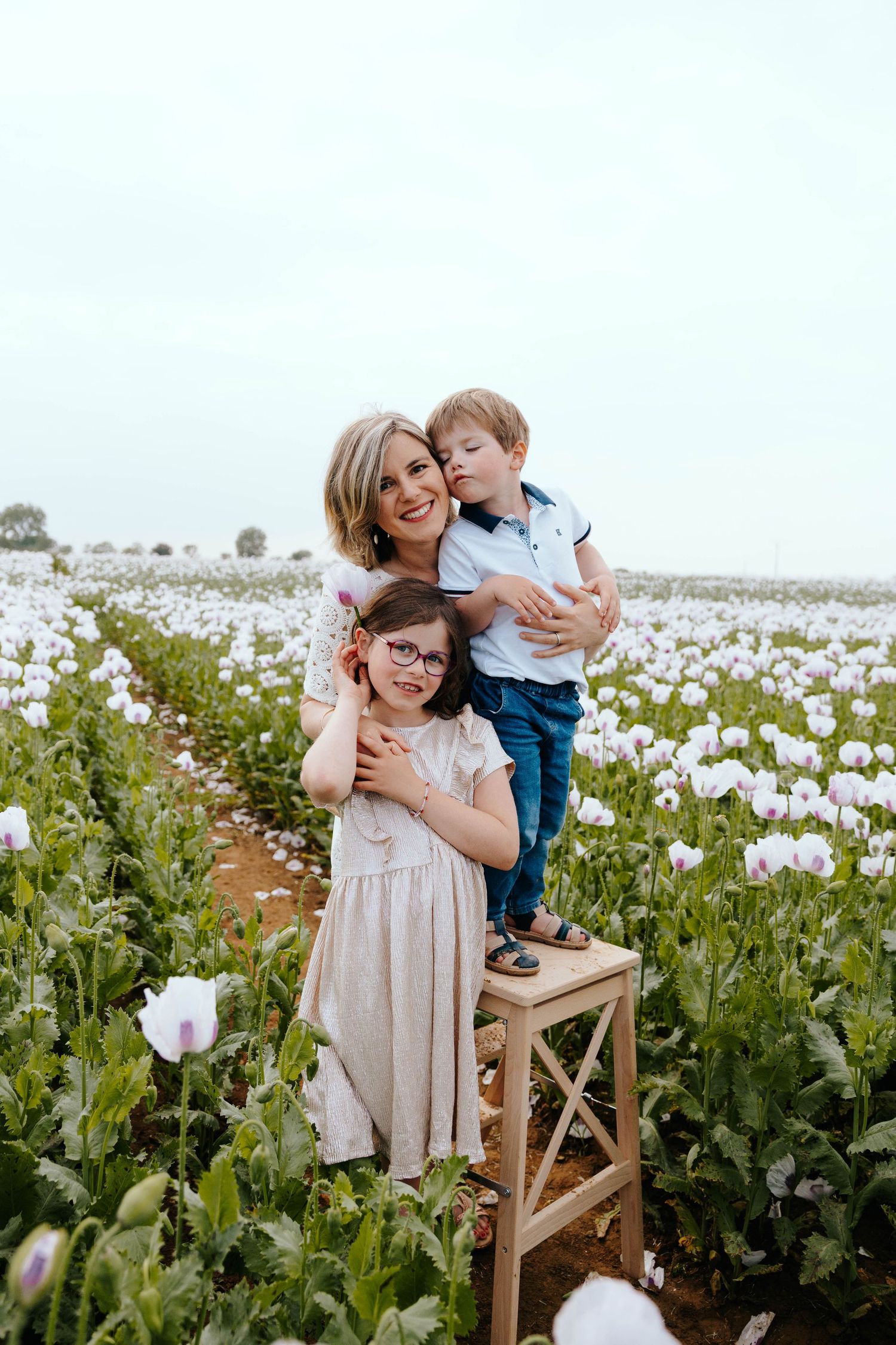 Mother hugging daughter and son standing on wooden stool in a white poppy flower field.