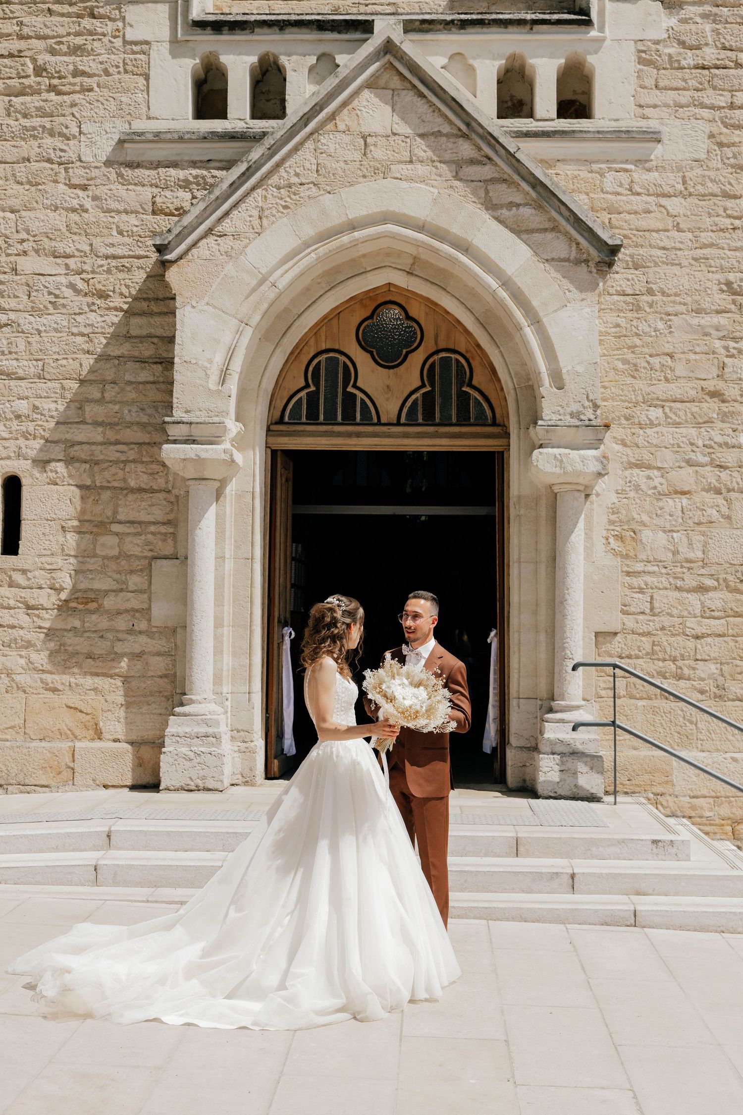 Bride and groom stand before a stunning Gothic stone church entrance, bride holding a white floral bouquet.