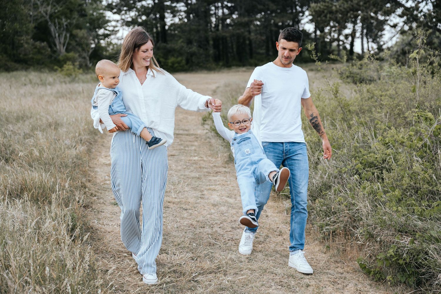 Joyful family of four walking on a nature path, parents swinging young boy while mother carries baby.
