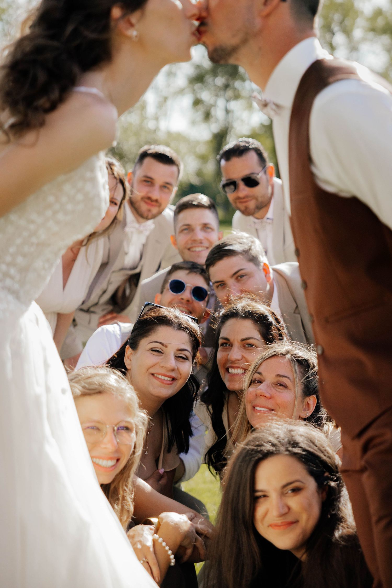 Wedding couple kissing above smiling group of friends posing together outdoors in sunlight.