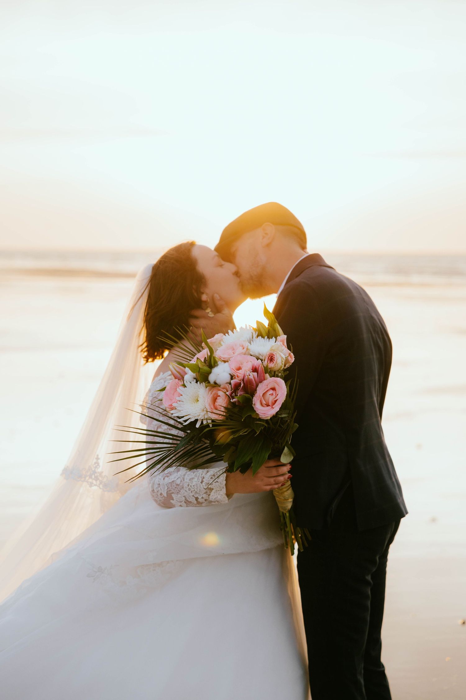 Bride and groom kissing on beach at sunset, bride holding pink floral bouquet with palm leaf, wearing white gown and veil.