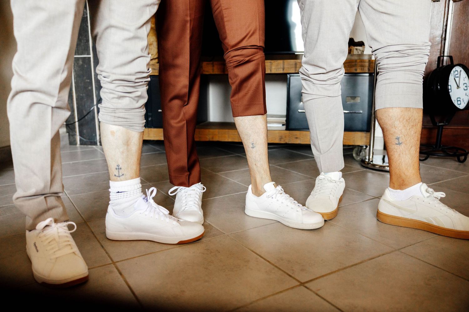 Four people wearing white sneakers and colorful pants standing together on tiled floor, some with ankle tattoos visible.