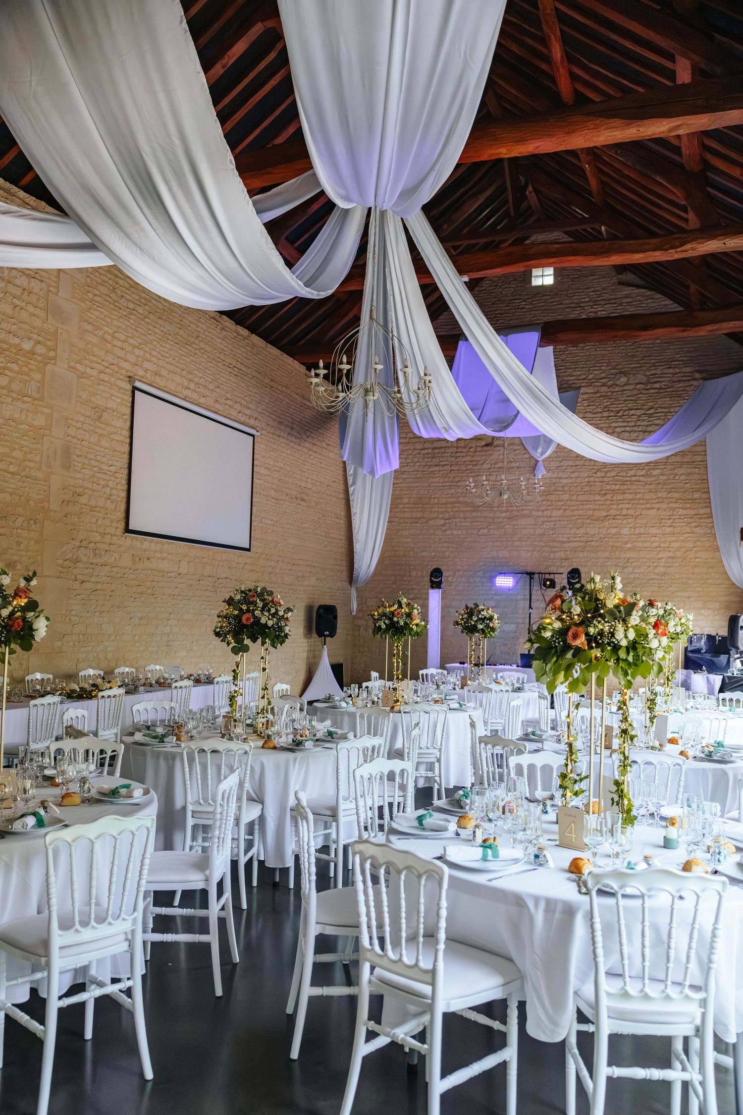 Wedding reception hall featuring white ceiling drapes, brick walls, projection screen, tall floral centerpieces, and white chairs.