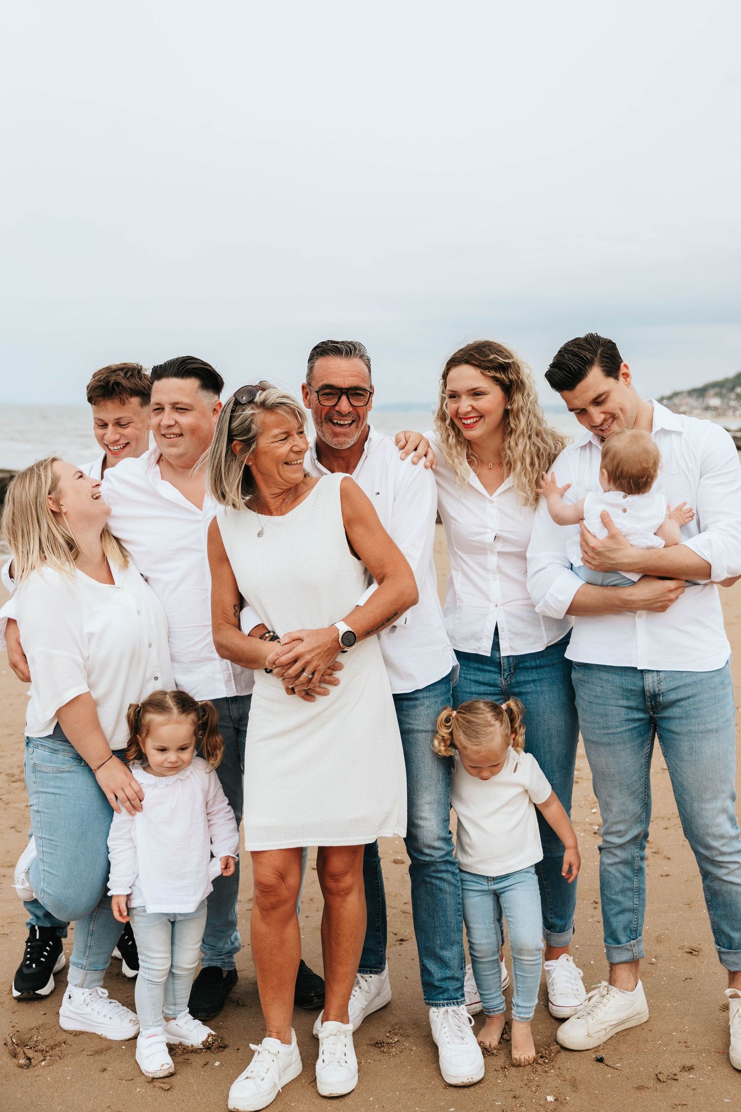 A large multigenerational family in white shirts and jeans poses together smiling on a sandy beach.